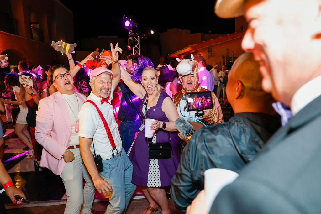 People dancing and having fun at a lively party with colorful lights, some wearing costumes like a cow and a pig, and a person being filmed by someone with a smartphone.