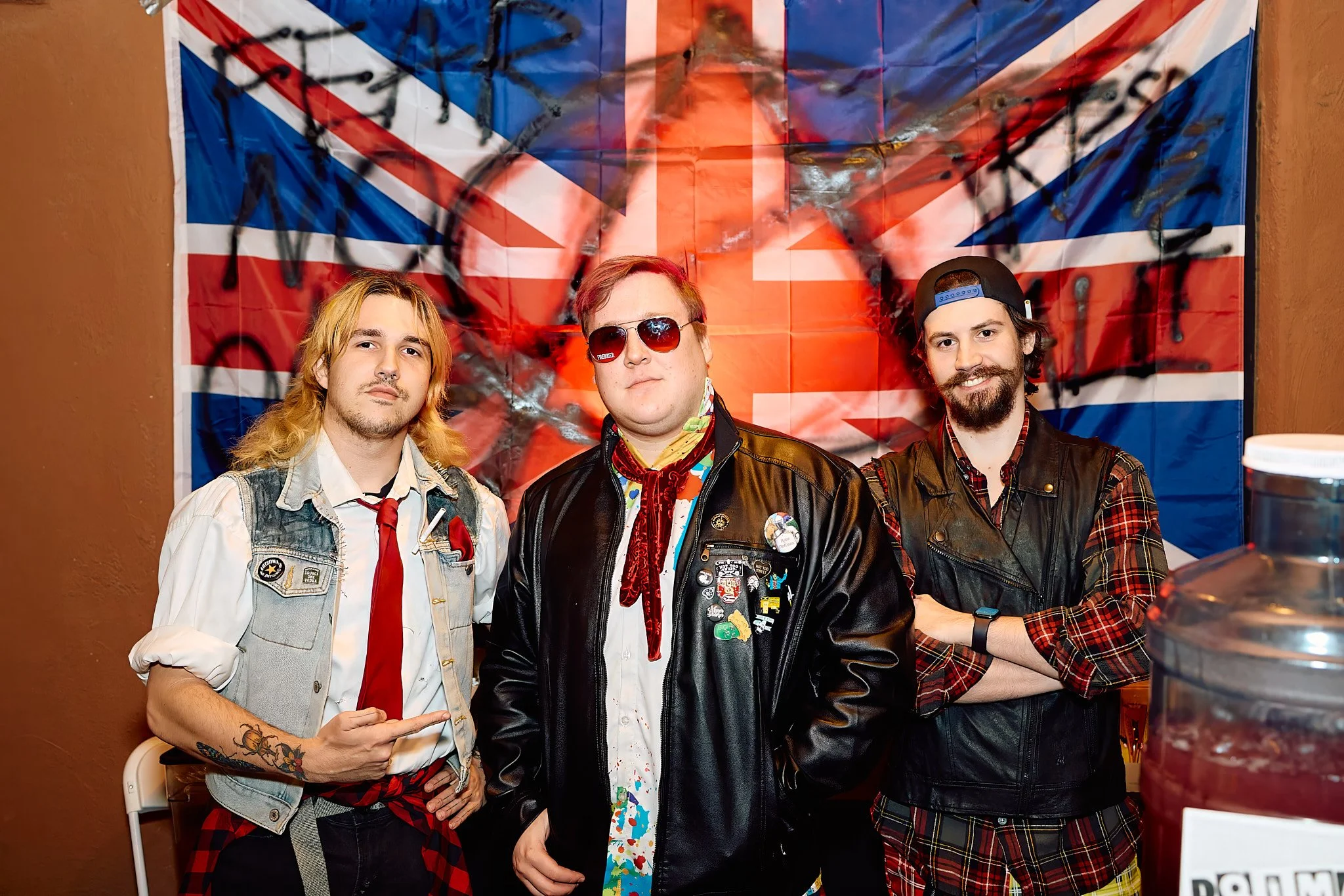 Three young men standing in front of a red, white, and blue Union Jack flag with black spray paint markings. The man on the left has long blonde hair, tattoos, and is wearing a denim vest with patches and a white shirt, pointing with his right hand. The man in the middle is wearing sunglasses, a black leather jacket with pins, and a colorful shirt underneath. The man on the right has a beard, a backward cap, a plaid shirt, and a black leather vest. They are all posing for the photo indoors.