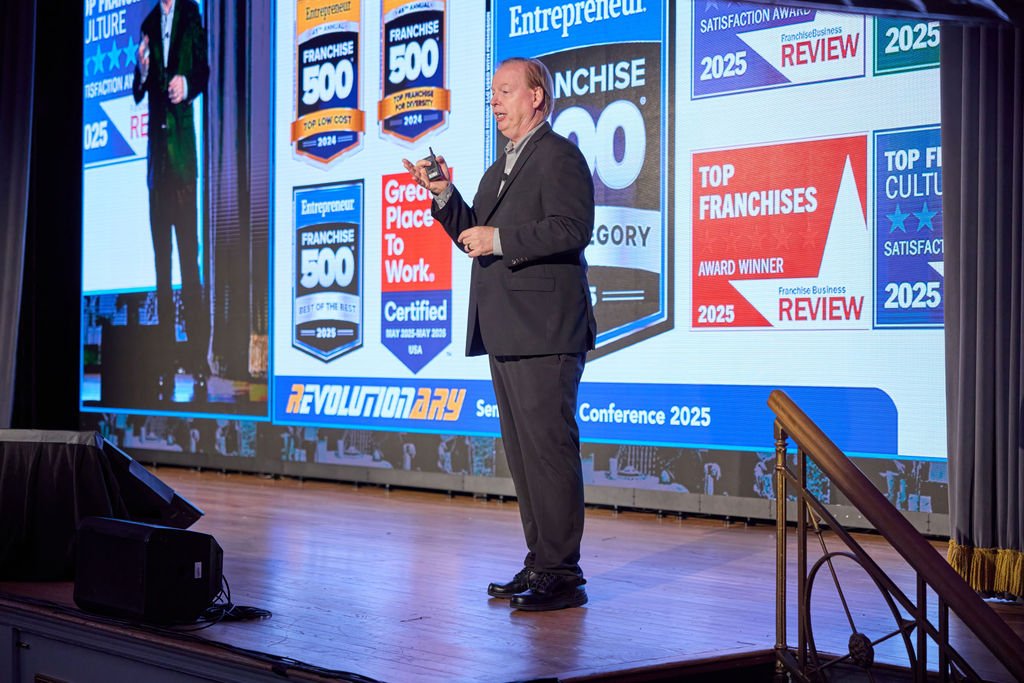 Man in a black suit speaking at a conference with large screen behind him displaying awards and badges for franchise recognition in 2024 and 2025.