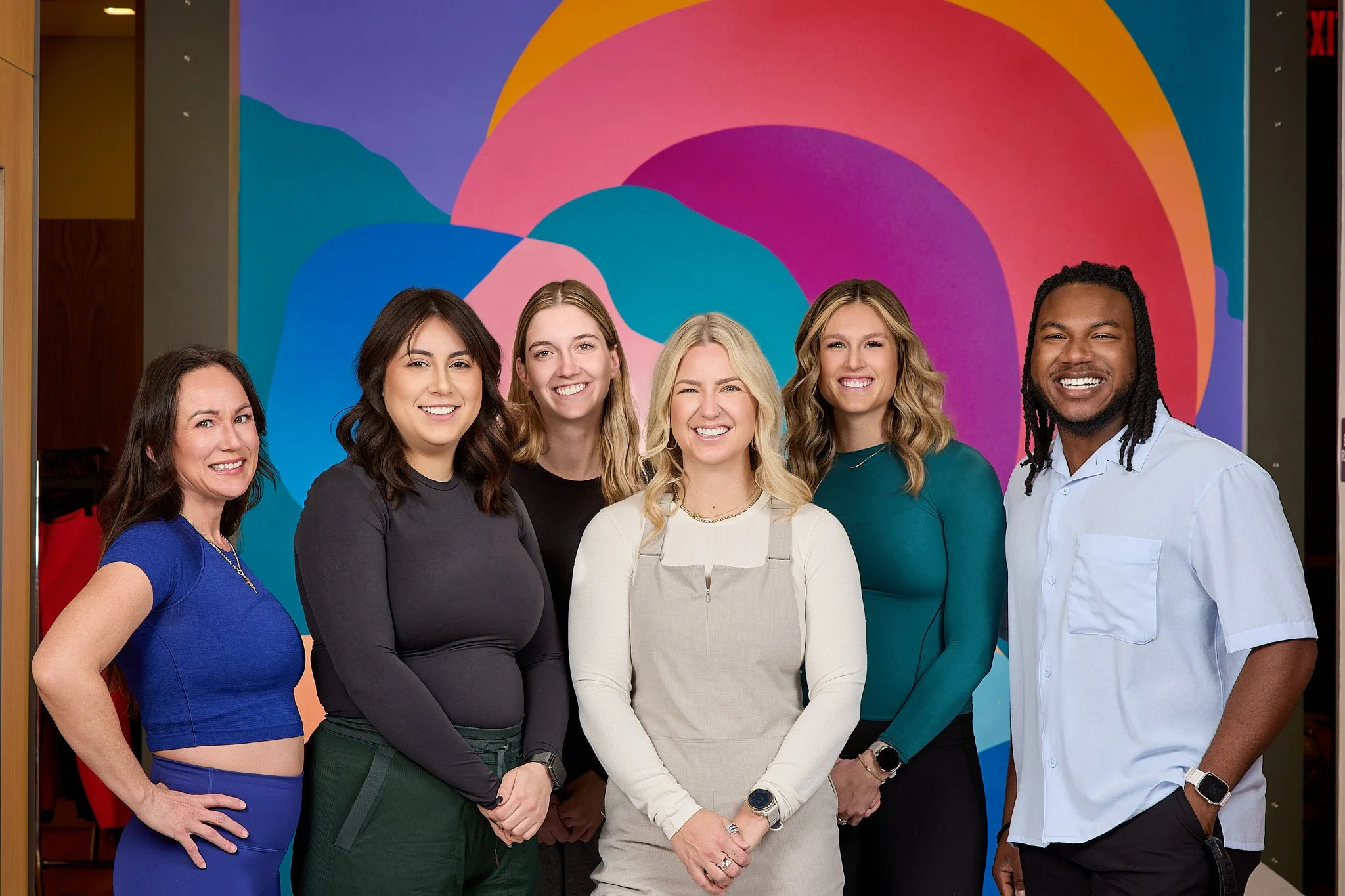 A group of six diverse women and one man standing in front of a colorful abstract mural, smiling and posing for a photo.