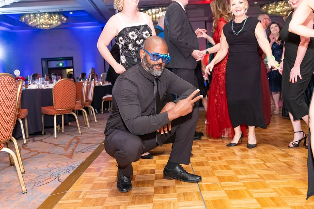 A man wearing black clothes and blue futuristic sunglasses squats on the dance floor at a formal party, making a peace sign with his right hand. He is surrounded by women in elegant dresses, dancing and holding hands in a decorated ballroom with tabl