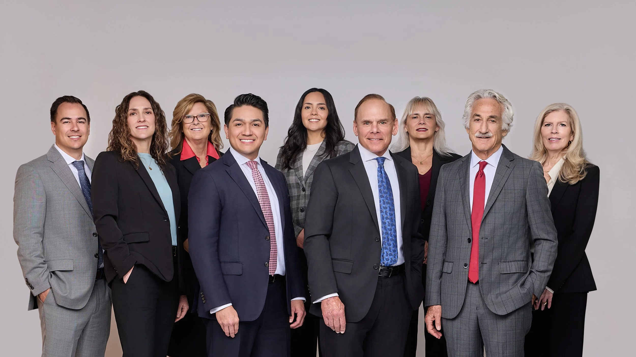 A diverse group of ten professionally dressed men and women standing together against a plain gray background, smiling for the camera.