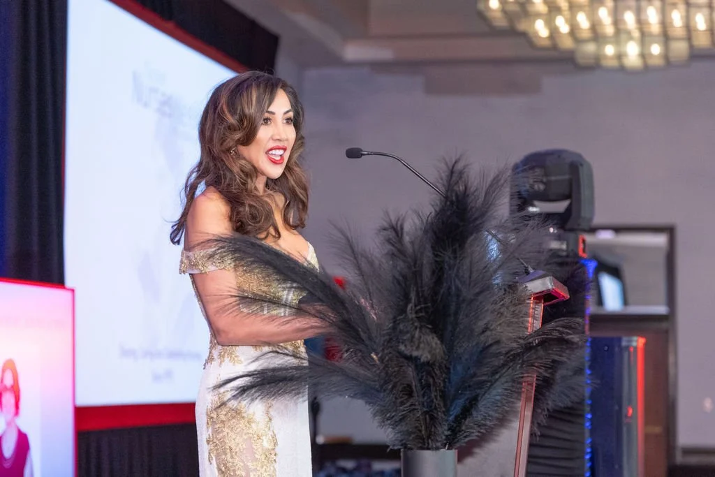 A woman giving a speech at a podium with a microphone, decorated in elegant black feathers, in a formal setting with a large screen and audio equipment in the background.