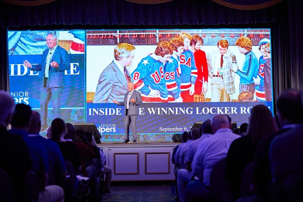 A speaker on stage at a conference presenting a slide titled 'Inside the Winning Locker Room,' featuring a large photo of a hockey team and a smaller photo of a man in a suit speaking at a podium. The audience is seated facing the stage.