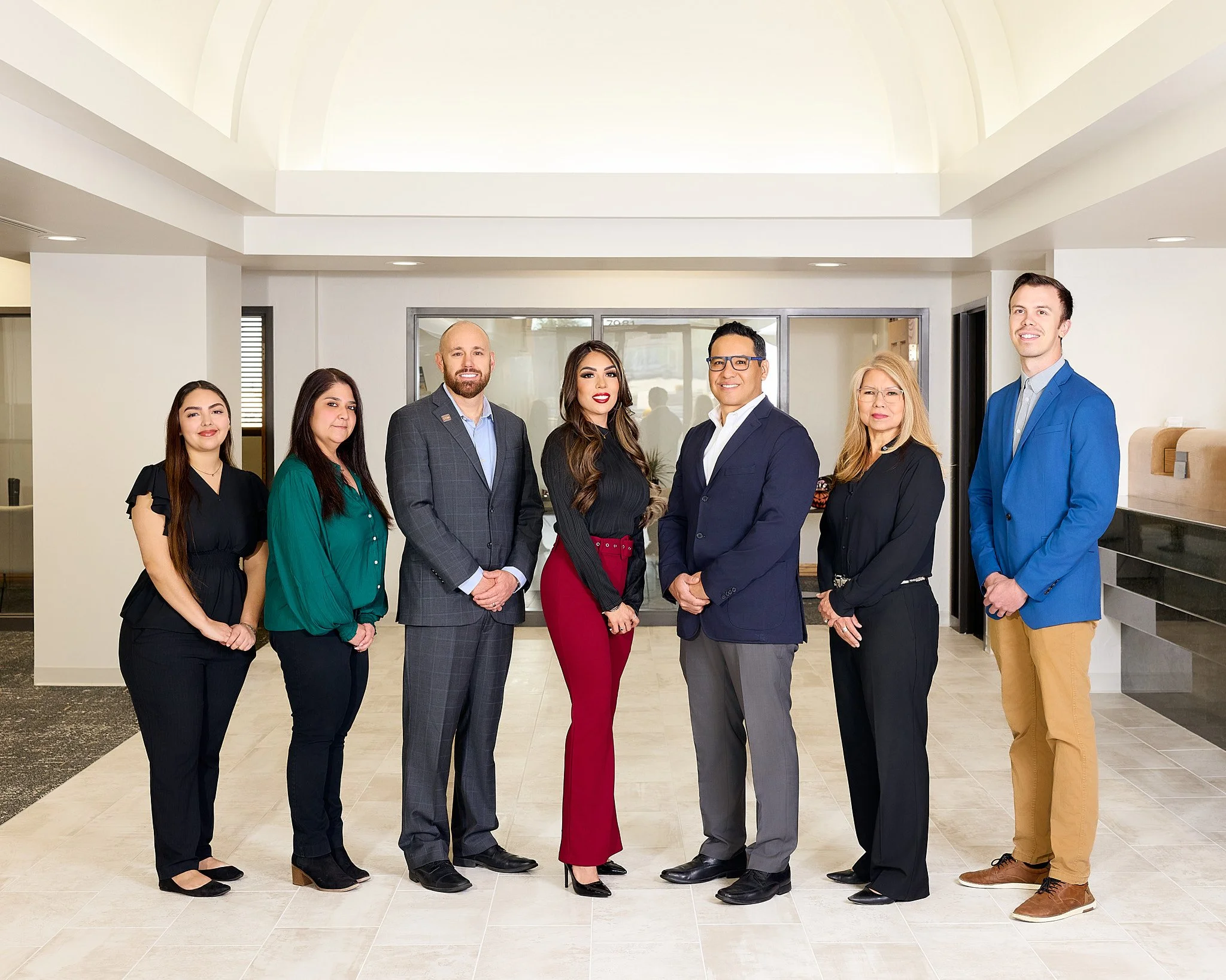 A diverse group of seven professionals standing together indoors in front of a glass door, dressed in business attire, smiling and looking at the camera.