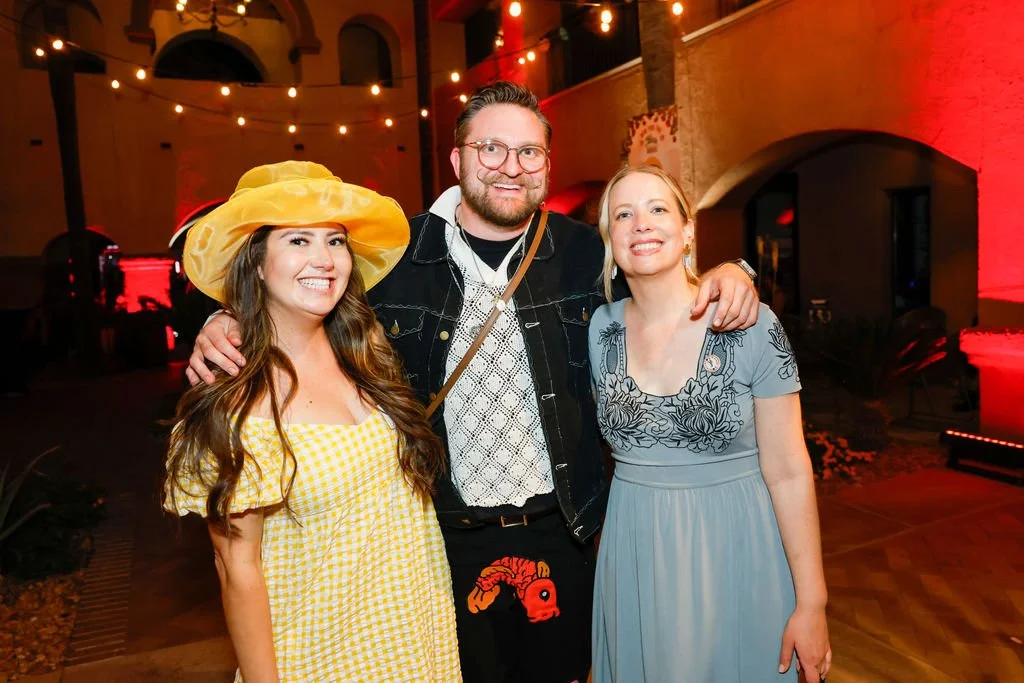 Three people smiling at a party or gathering, two women and one man, with string lights hanging overhead and colorful lighting in the background.