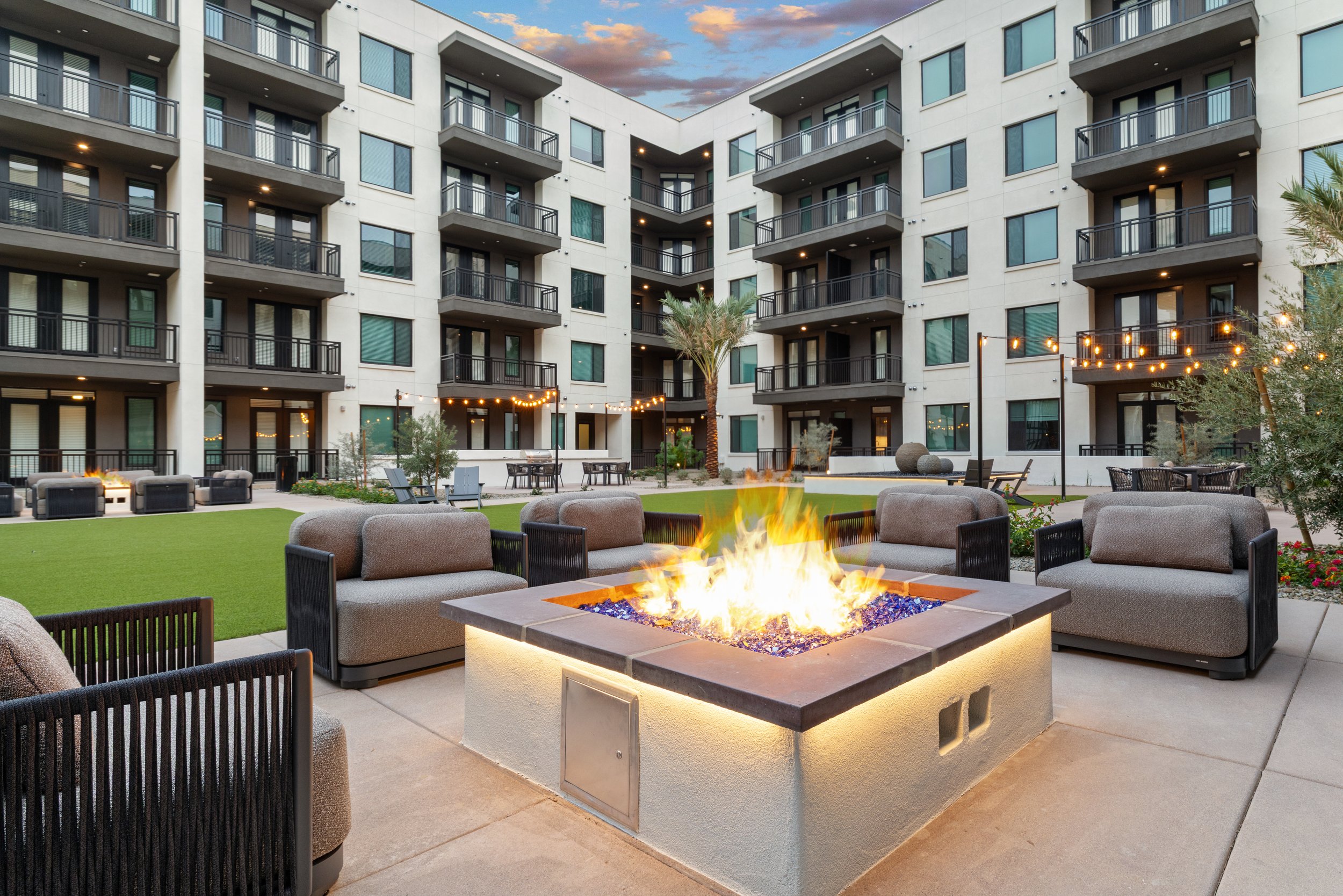 A courtyard in an apartment complex with outdoor seating, string lights, and a fire pit under a purple sky with clouds.