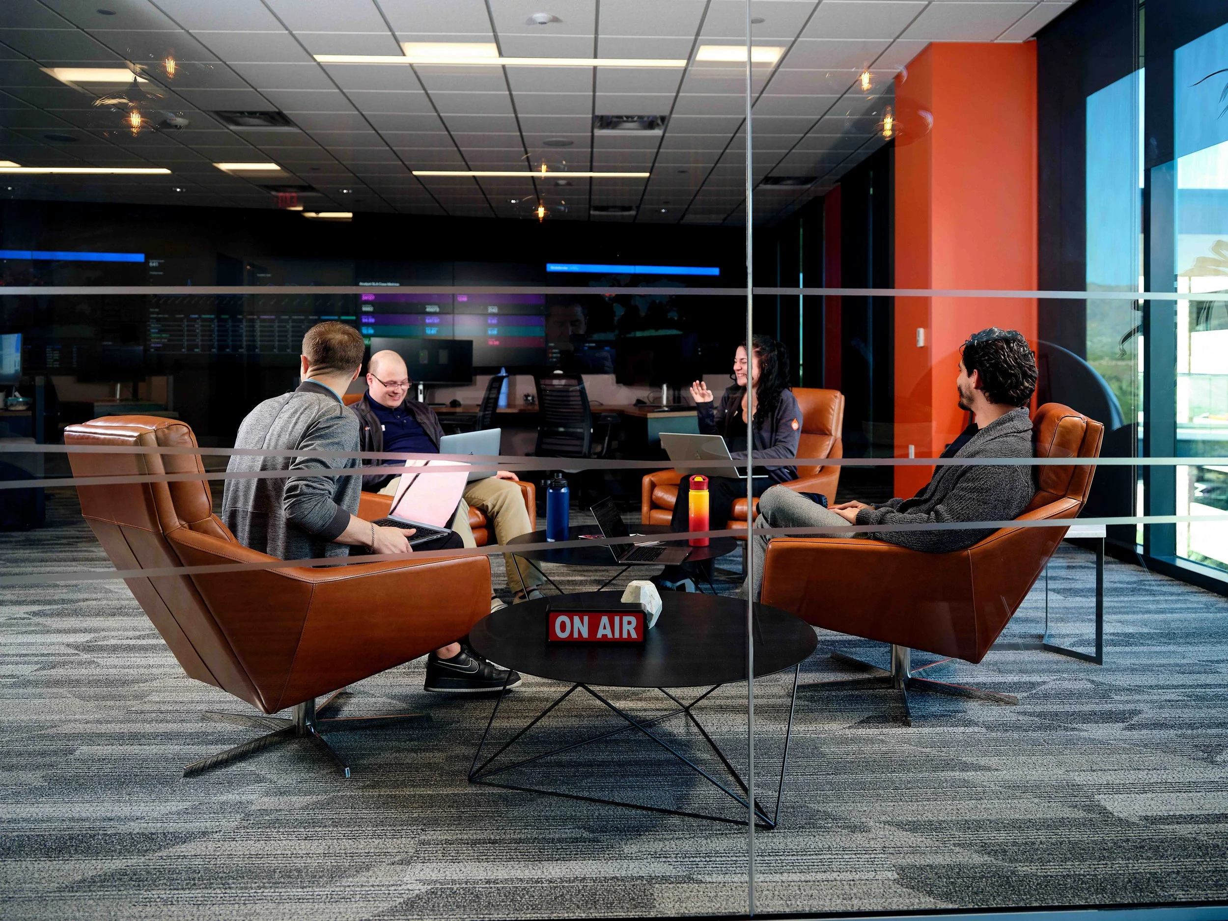 A group of four people sitting in leather chairs around a small circular table with an 'On Air' sign, inside a modern office with glass walls, orange and dark colored walls, and a digital display in the background.