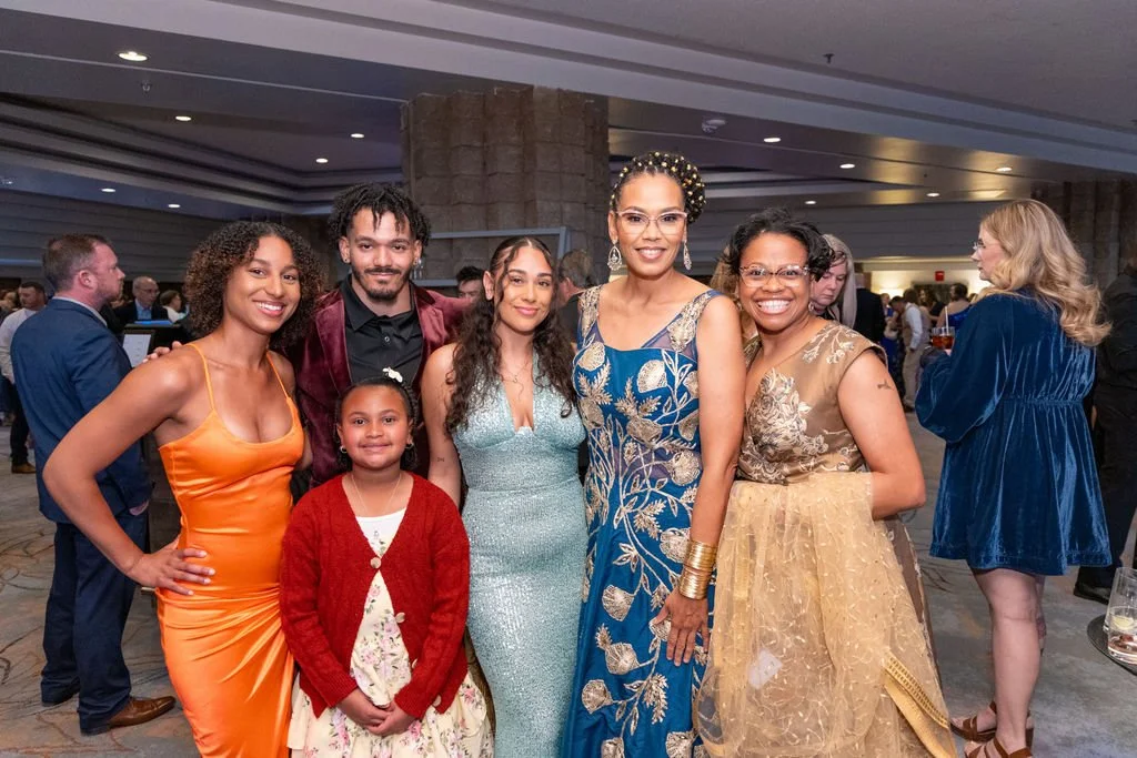 Group of six women and one young girl at a formal event, posing for a photo, with a busy background of other guests.