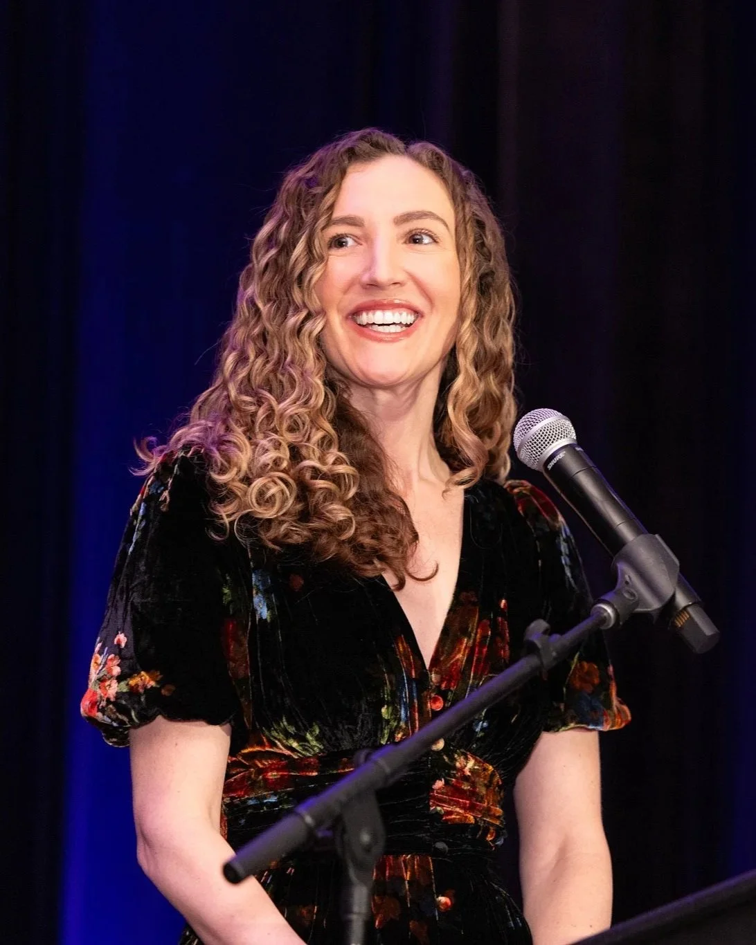 A woman with curly hair smiling while speaking in front of a microphone on stage.