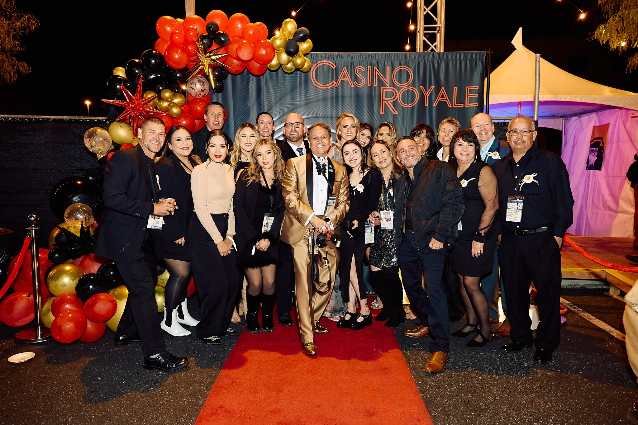 A group of people dressed in formal attire posing on a red carpet at a Casino Royale event, with balloons and a decorated background.