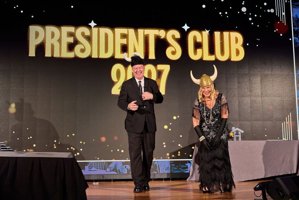 Two people dressed in costumes on stage at a 2025 President's Club event, with a large screen behind them displaying the event name.