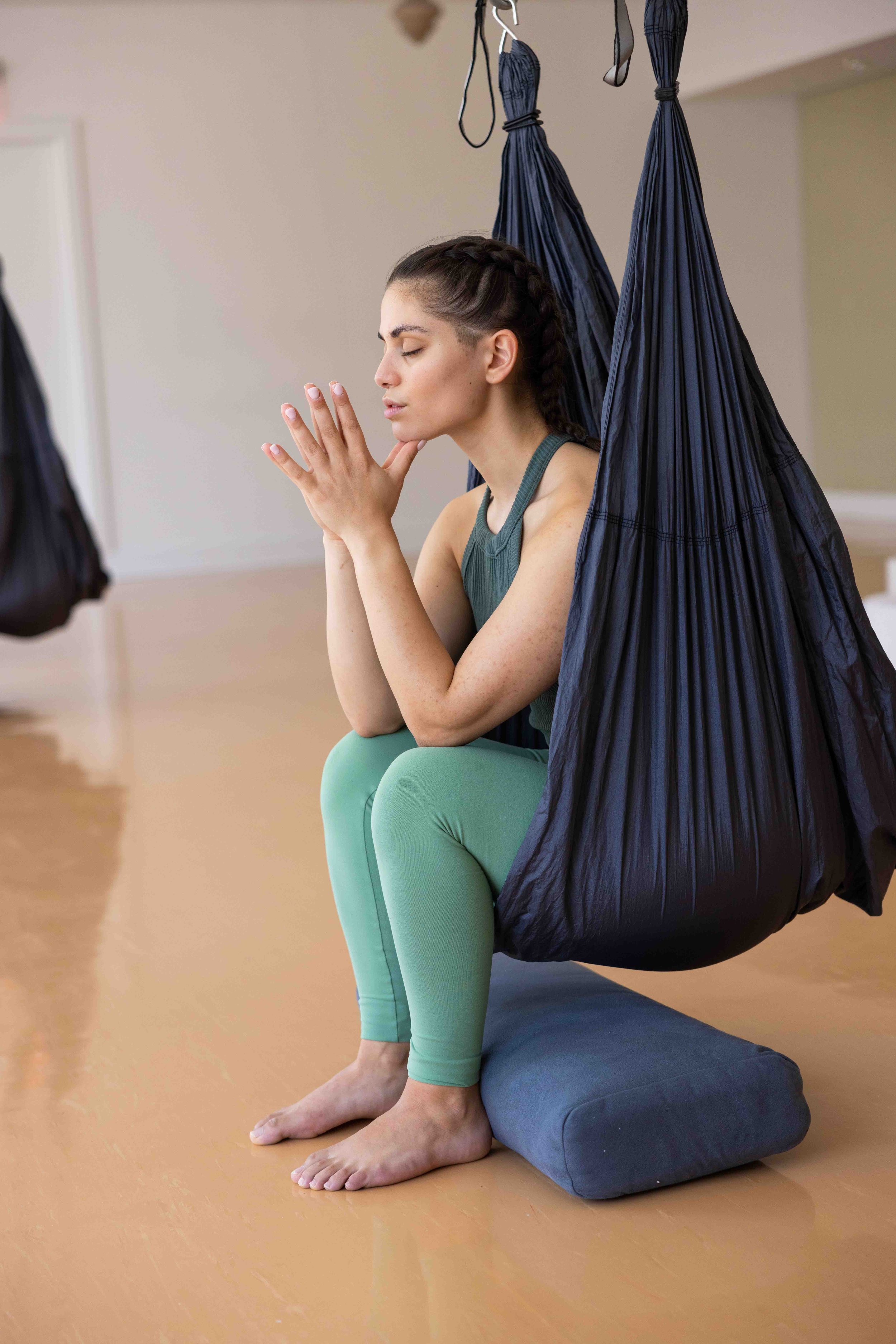 A woman practicing yoga or meditation in a yoga hammock, sitting cross-legged with hands together near her face, eyes closed, in a well-lit room with light-colored walls and wooden flooring.