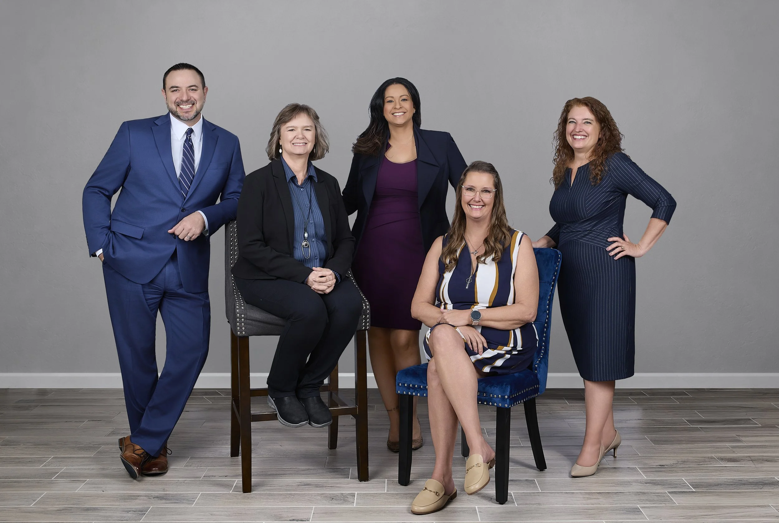 Six diverse professionally dressed individuals, four women and two men, posing in a room with a plain gray wall and wood flooring. The group appears confident and friendly, with a mix of seated and standing positions.