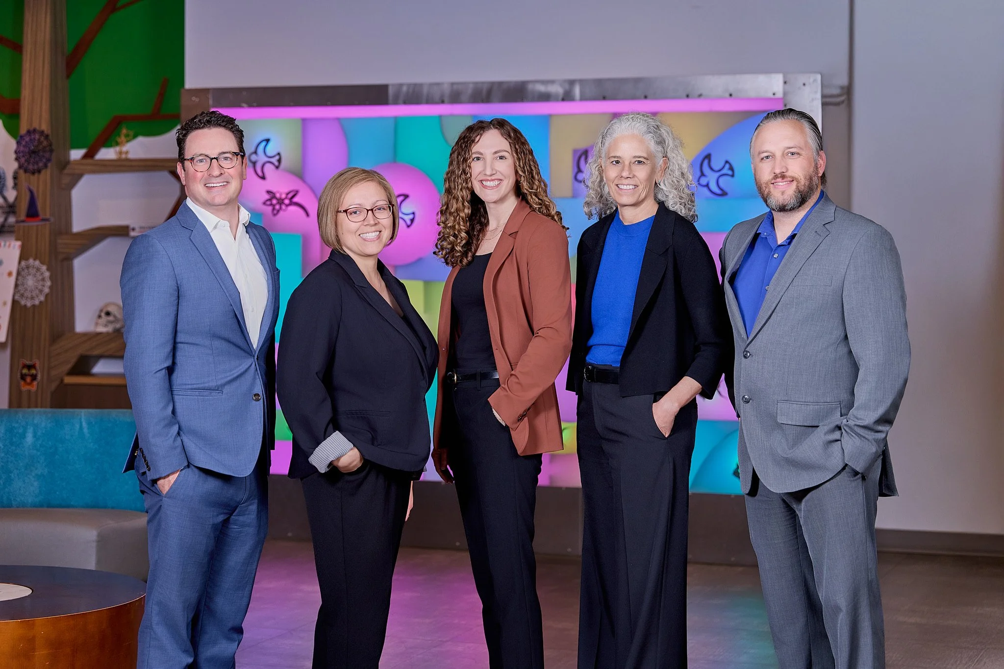 Five professionally dressed people standing together, smiling, indoors with colorful abstract background.