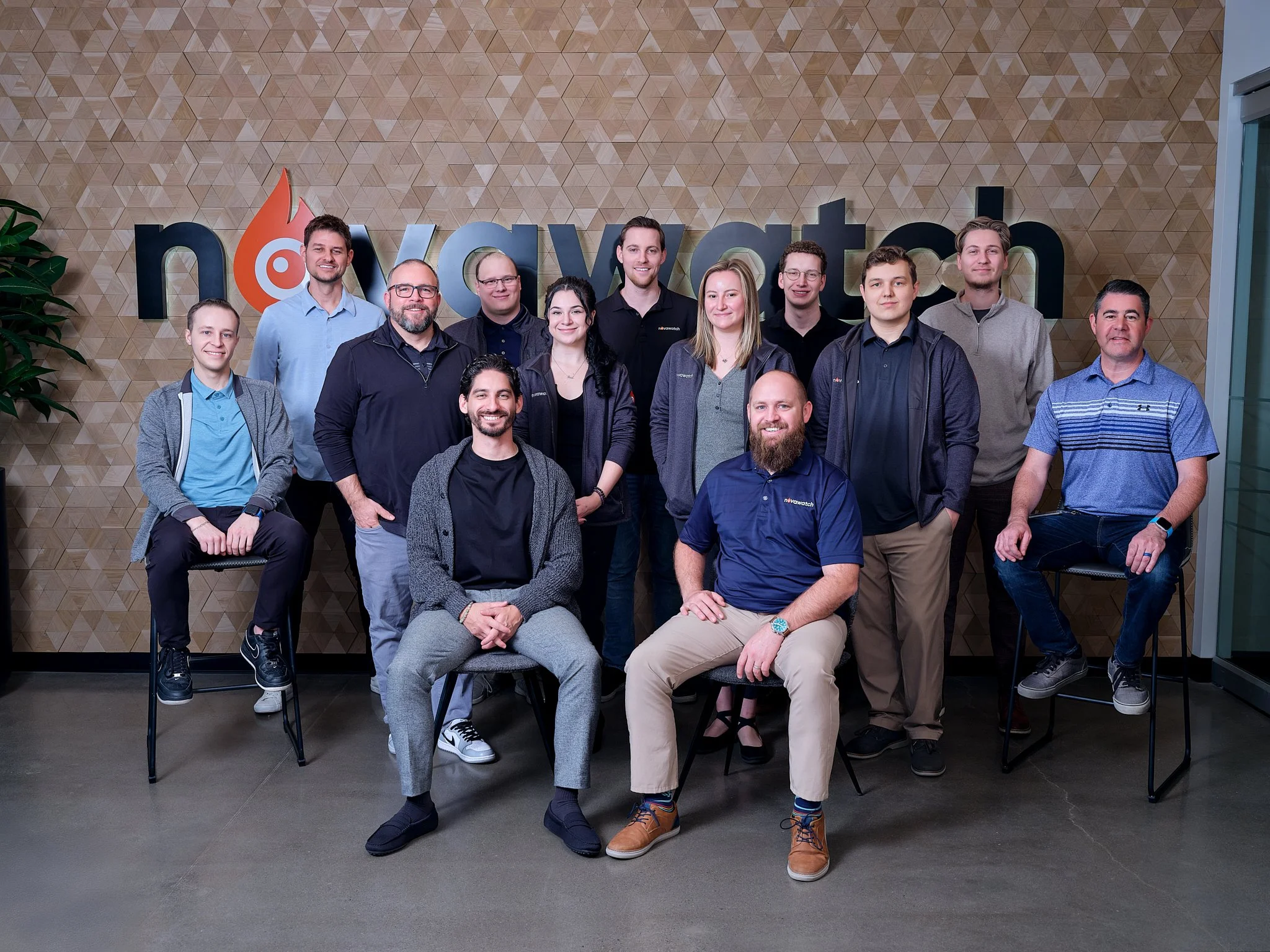 A group of fourteen diverse people posing indoors in front of a wooden textured wall with a partially visible logo that reads 'nova watch'. They are standing and sitting in two rows, smiling at the camera.