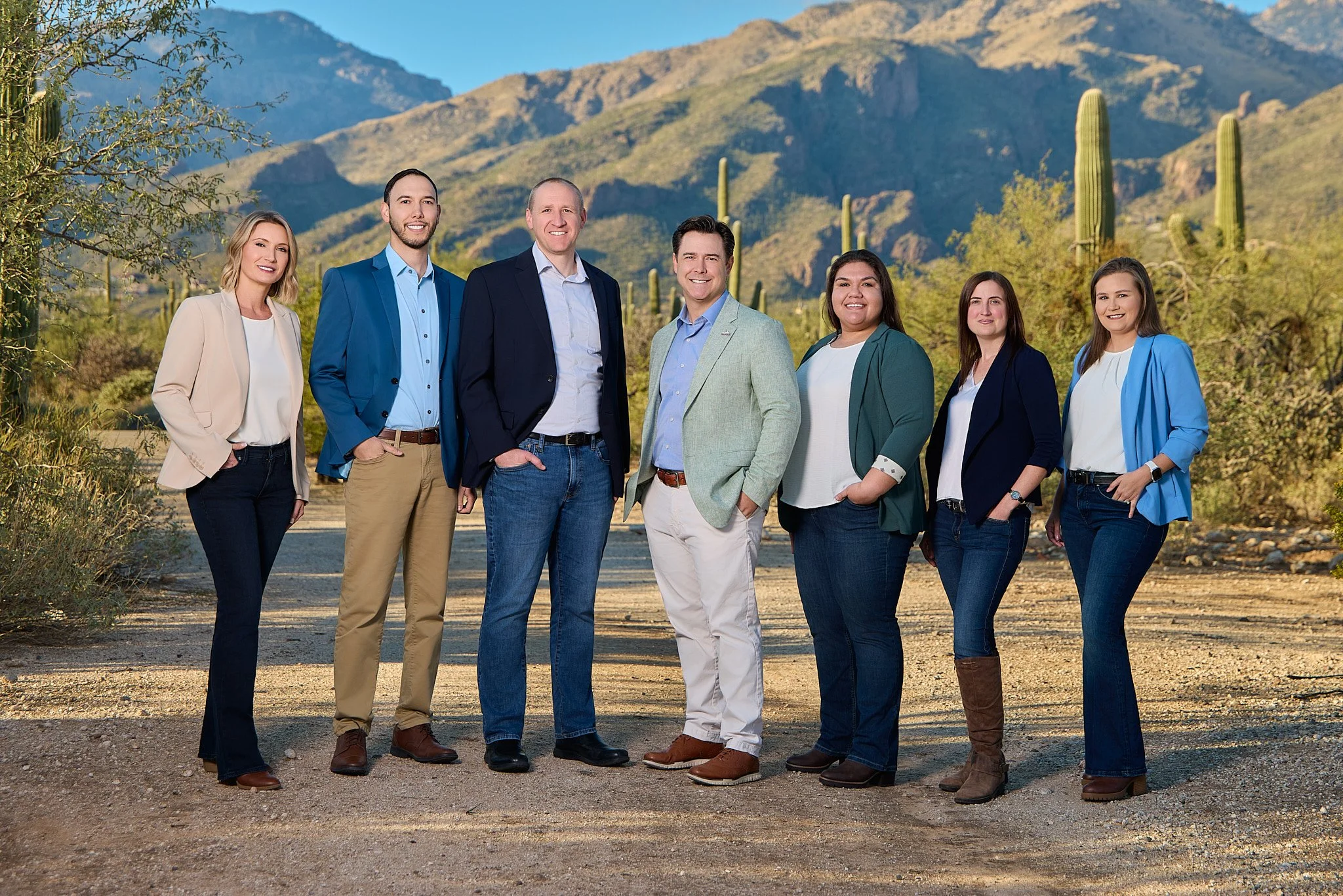 A diverse group of seven people, five women and two men, standing outdoors on a dirt path with mountains, flowering plants, and tall cacti in the background, dressed in business casual attire, smiling at the camera.