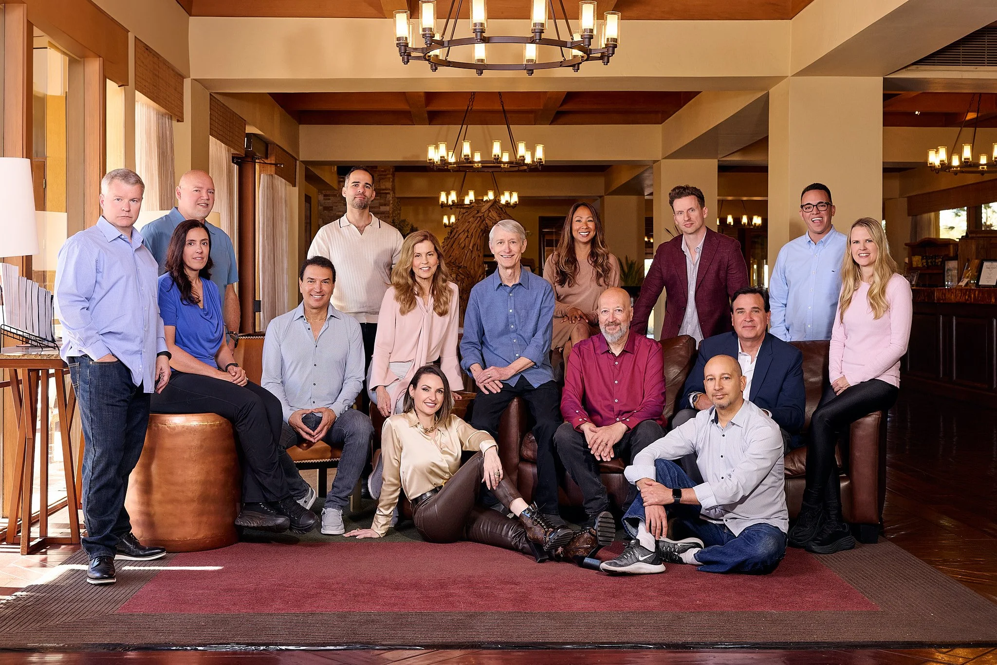 A group of fifteen diverse people gathered together for a photo in a warmly lit lobby or lounge area. Some are sitting on a couch, others standing behind. They are casually dressed and smiling at the camera.
