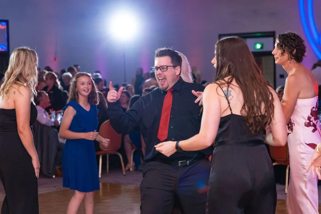 People dancing and enjoying themselves at a social event or party with colorful lighting and seated guests in the background.