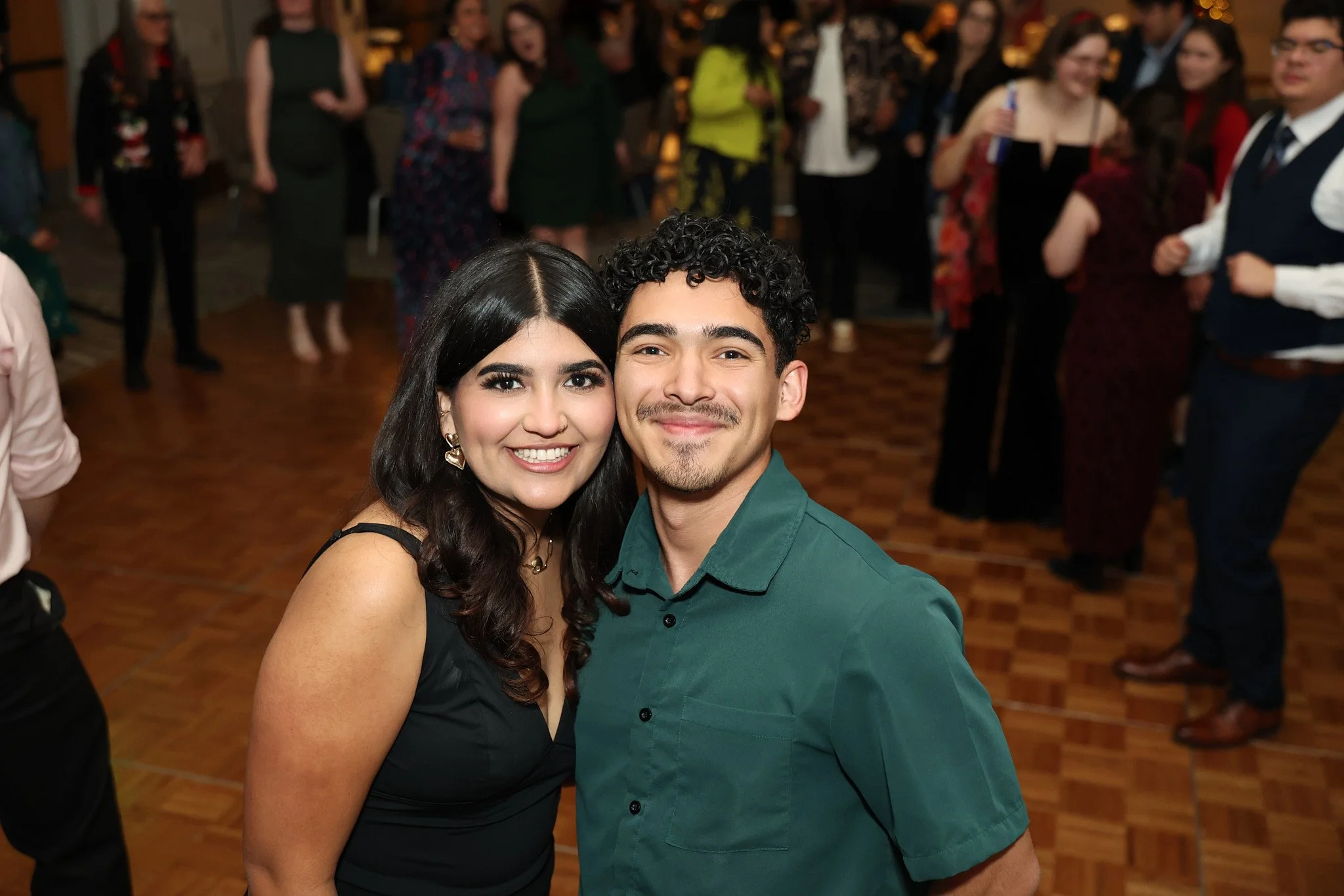 A smiling young woman with dark hair and earrings stands next to a smiling young man with curly hair, both dressed nicely, at a social event or party, with other people mingling and dancing in the background.