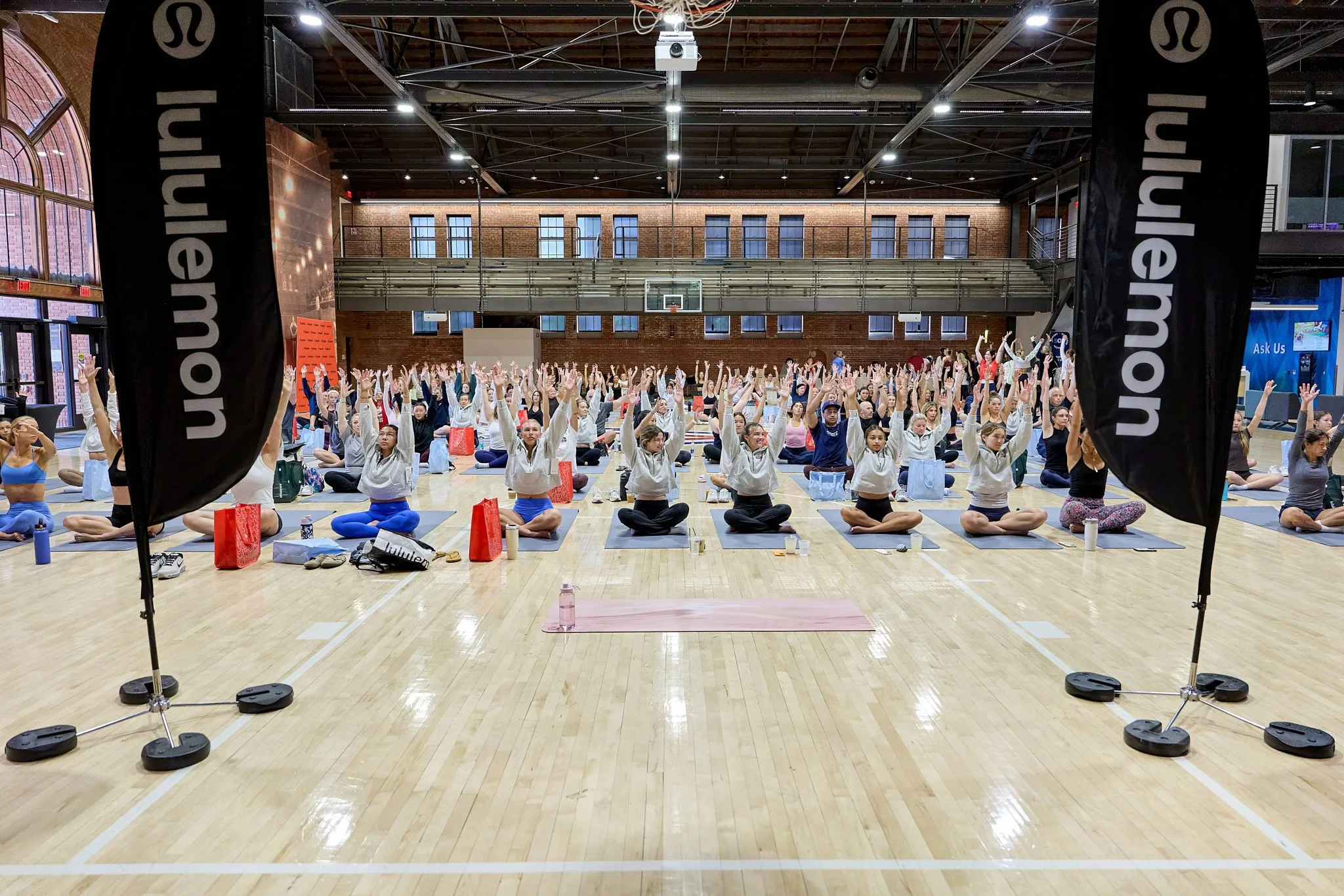 Large indoor yoga class with many participants in a gymnasium, seated on yoga mats with hands raised.