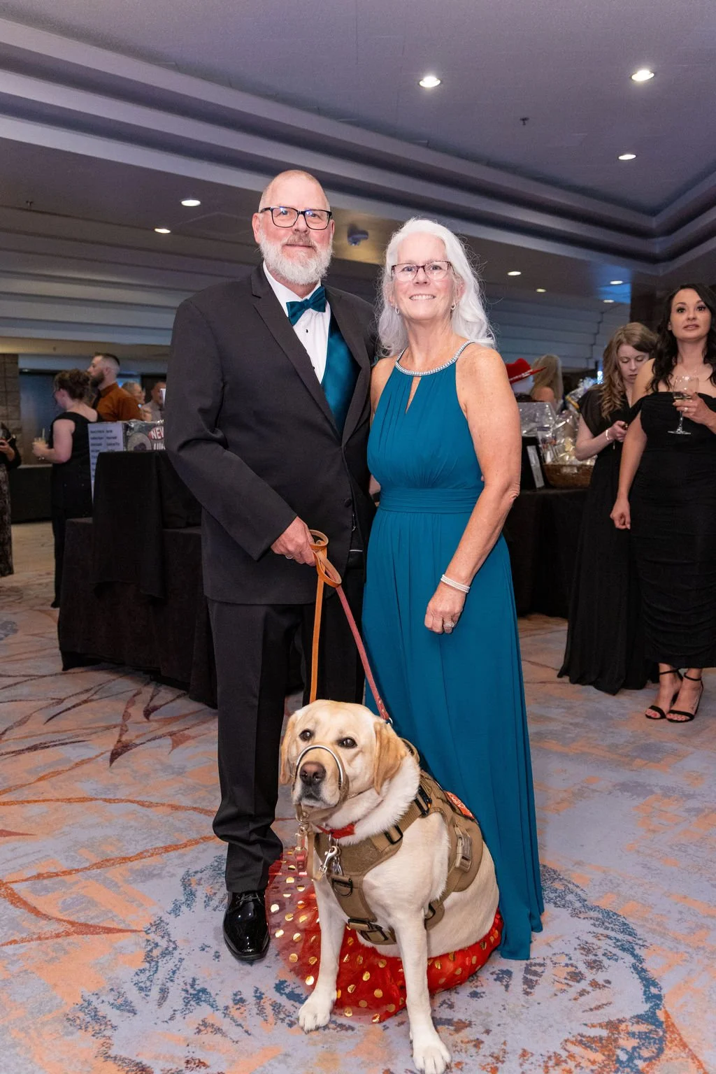 A man in a black tuxedo with a teal bow tie and a woman in a teal gown stand together at a formal event, holding a service dog sitting on a red polka-dotted mat. The man has glasses and a beard, while the woman has white hair and glasses. In the back