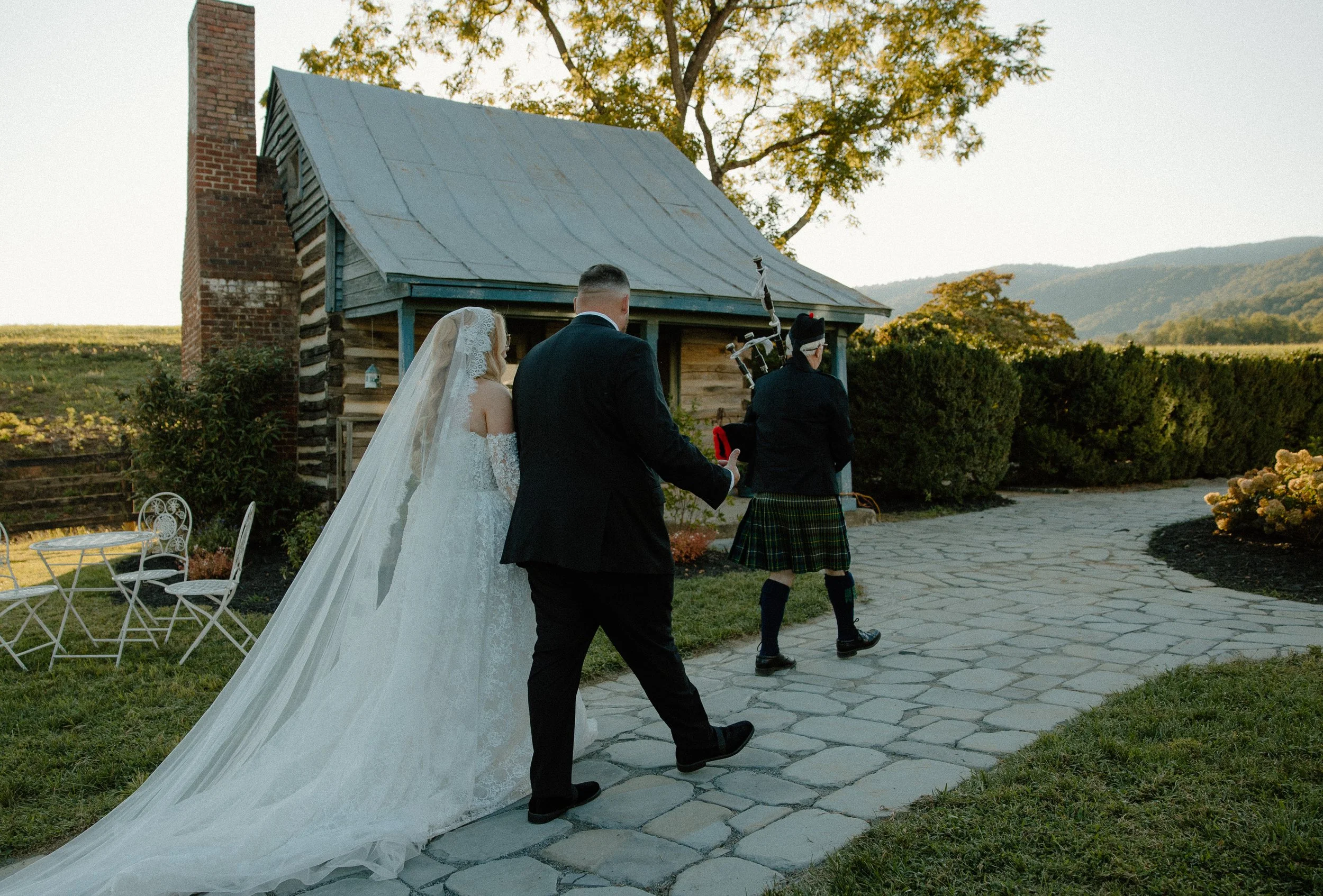 Bride and groom walking on a stone pathway outside an old wooden building with a metal roof, while a woman dressed in a kilt plays bagpipes in the background. The setting appears to be a rustic outdoor wedding or celebration during late afternoon or 