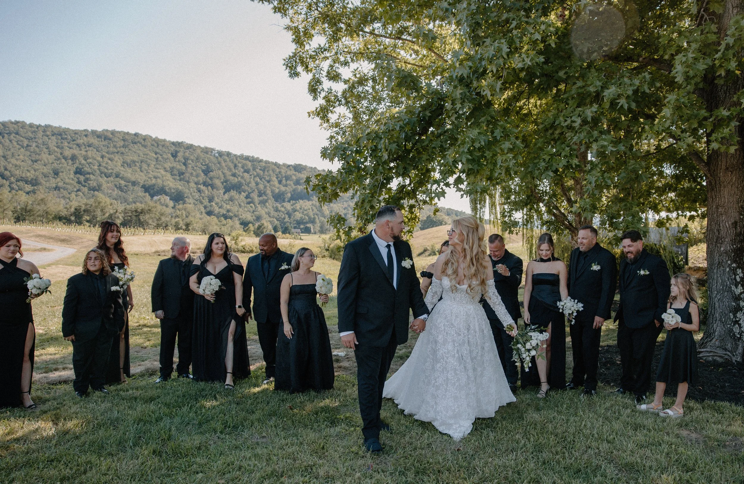 Bride and groom holding hands during an outdoor wedding ceremony in a grassy field under a large tree, with wedding party and guests in the background, surrounded by a scenic rural landscape with rolling hills and trees.