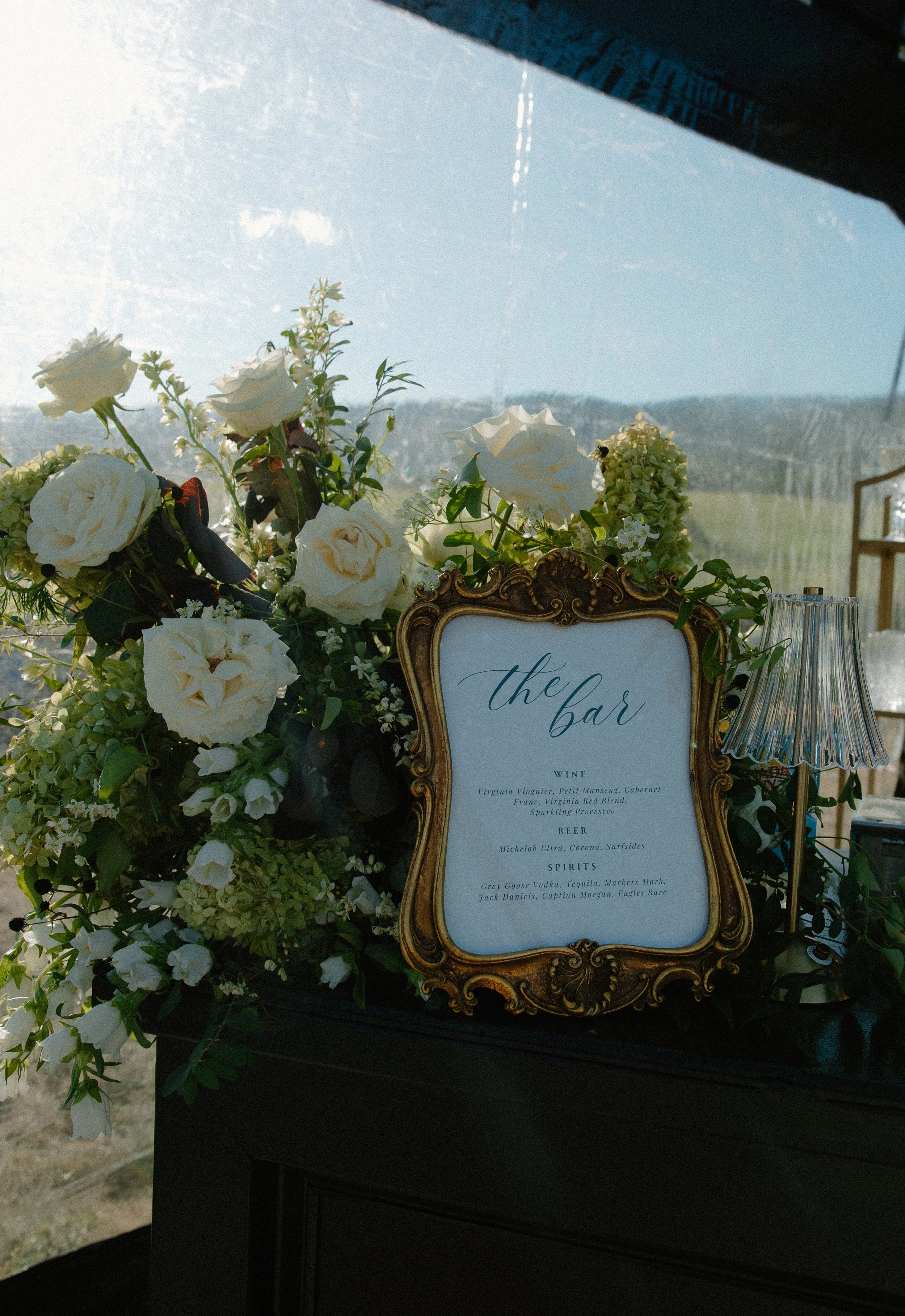 A floral arrangement with white roses and greenery next to a framed sign listing drinks at a bar, with a clear blue sky and distant fields in the background.