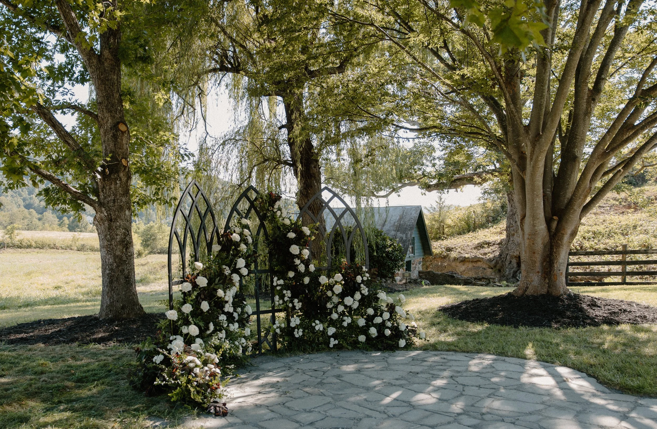 A garden scene with large trees, a black metal fence decorated with white flowers, a pathway, a small building with a sloped roof, and a field in the background.