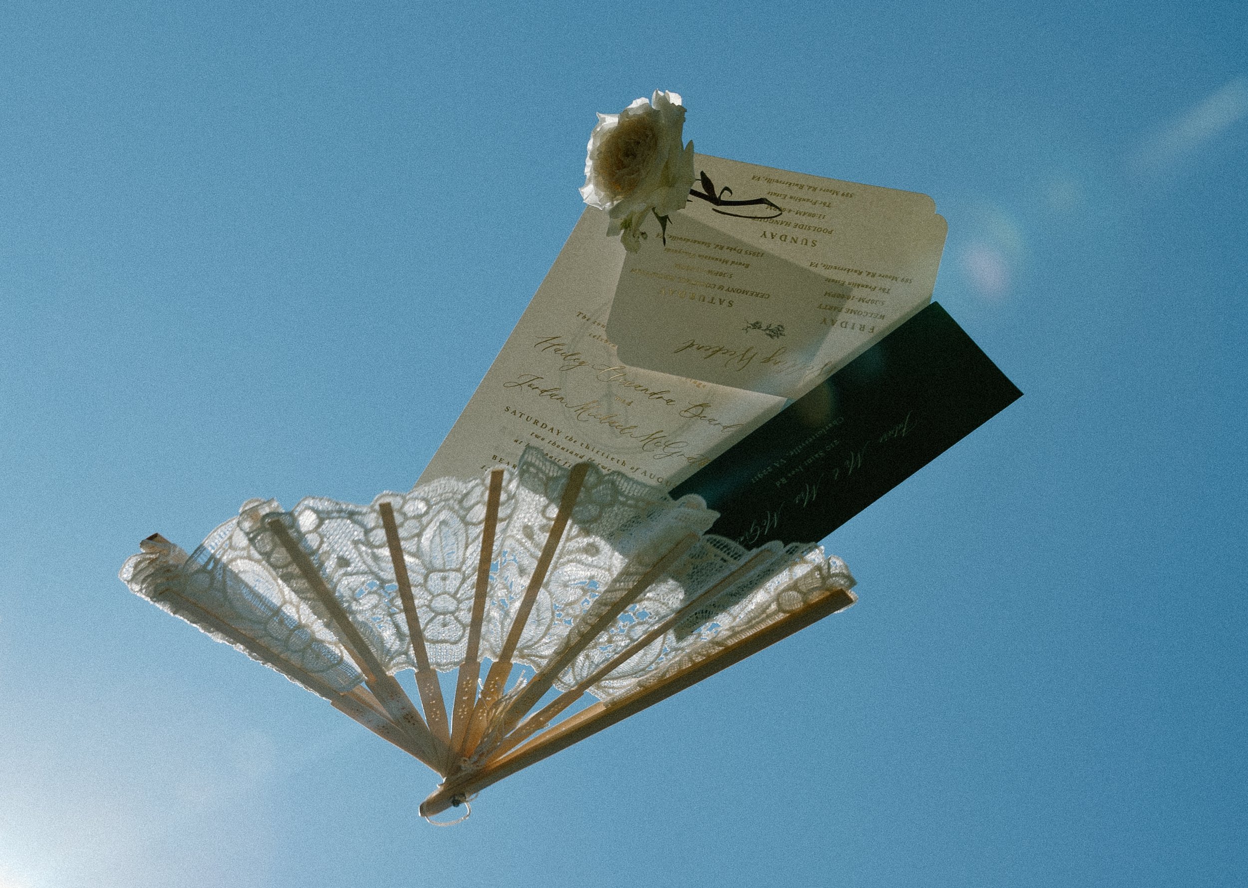 An ornate white handheld fan with lace details and a white flower attached, holding a wedding invitation, against a clear blue sky.
