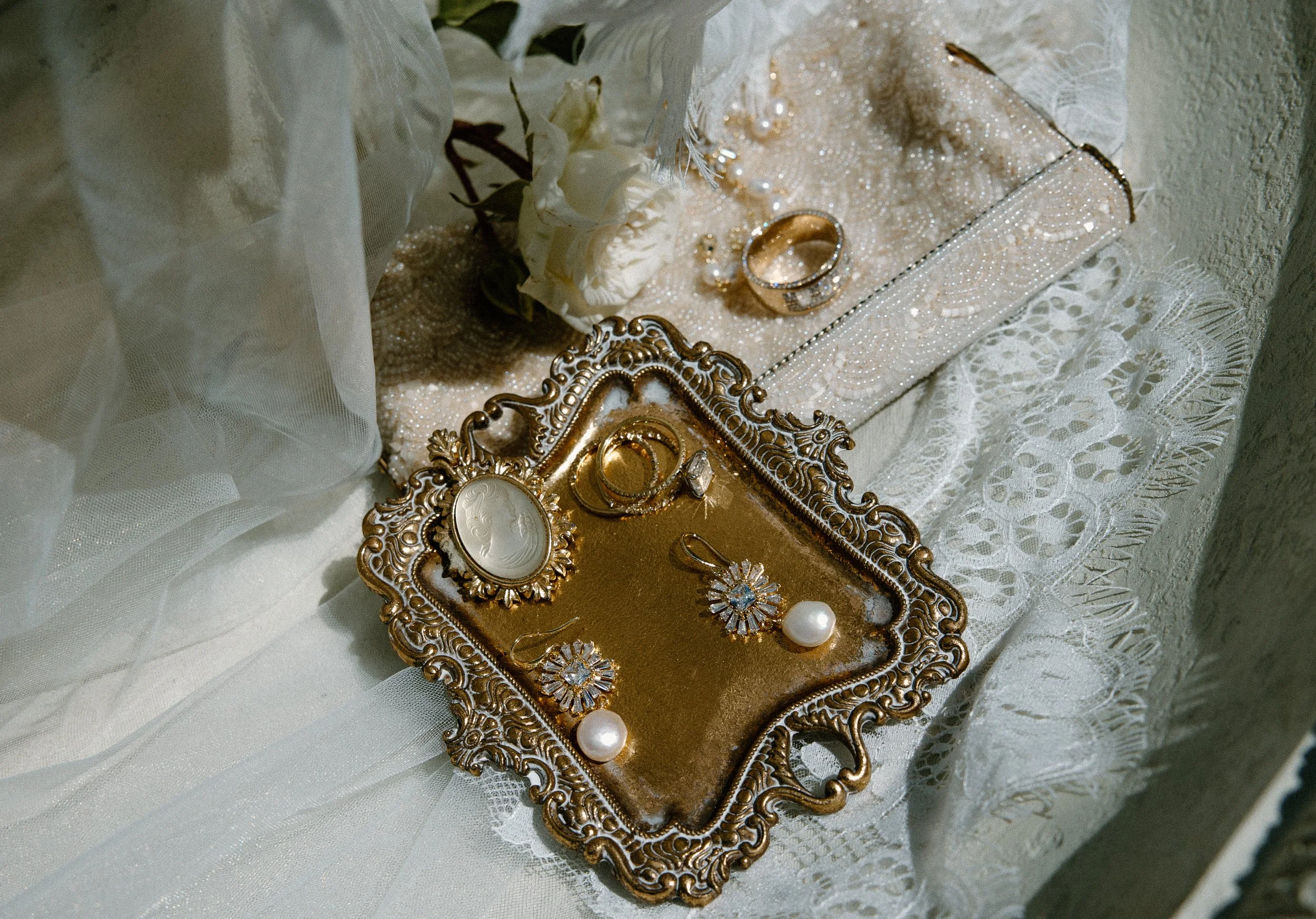 Jewelry display tray with rings, earrings, and a pearl, set on white lace fabric with white roses nearby.