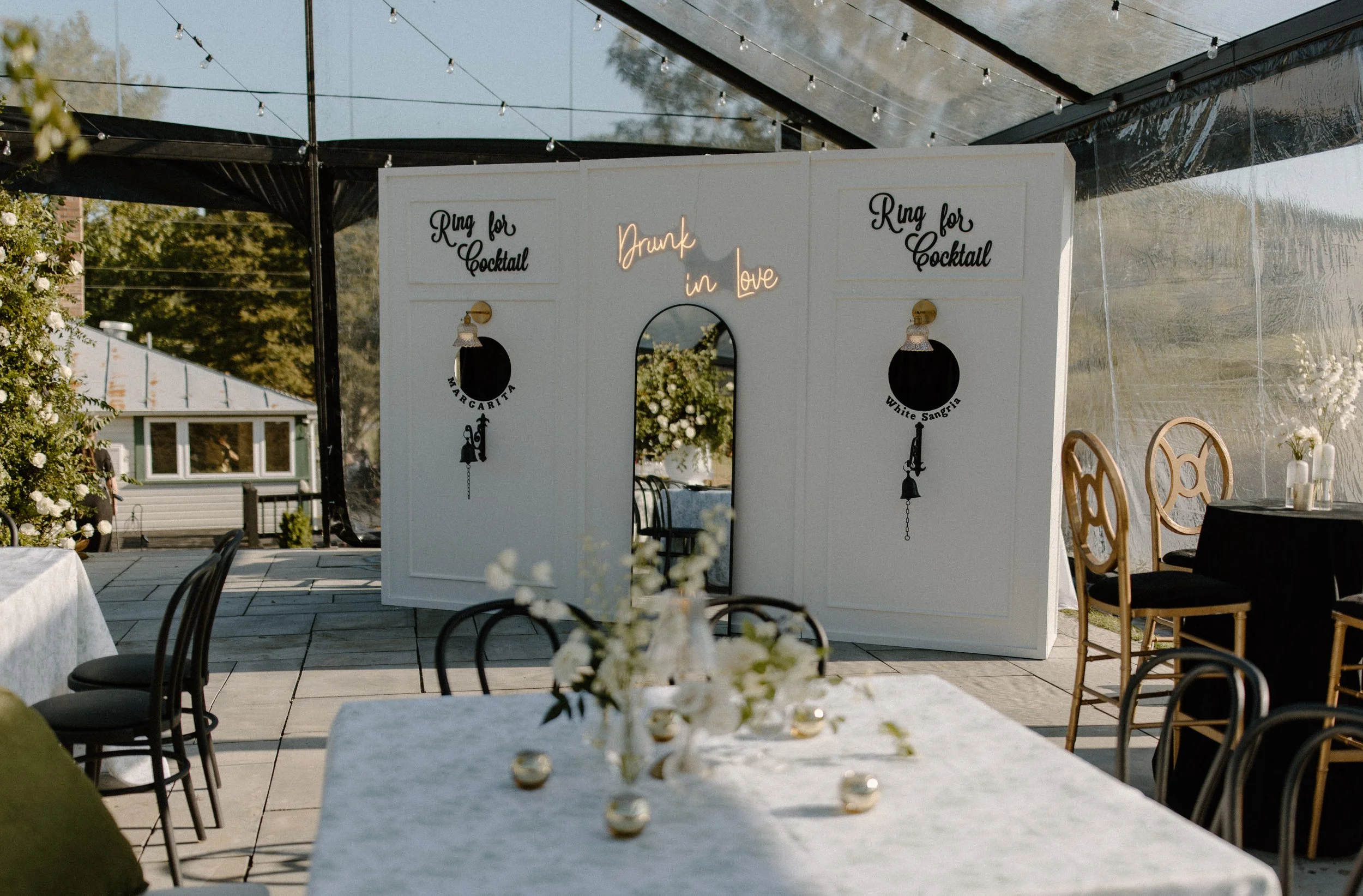 A decorated outdoor party area with a white backdrop featuring signs that say 'Ring for Cocktail' and 'Drink in love,' surrounded by string lights and tables with floral centerpieces.
