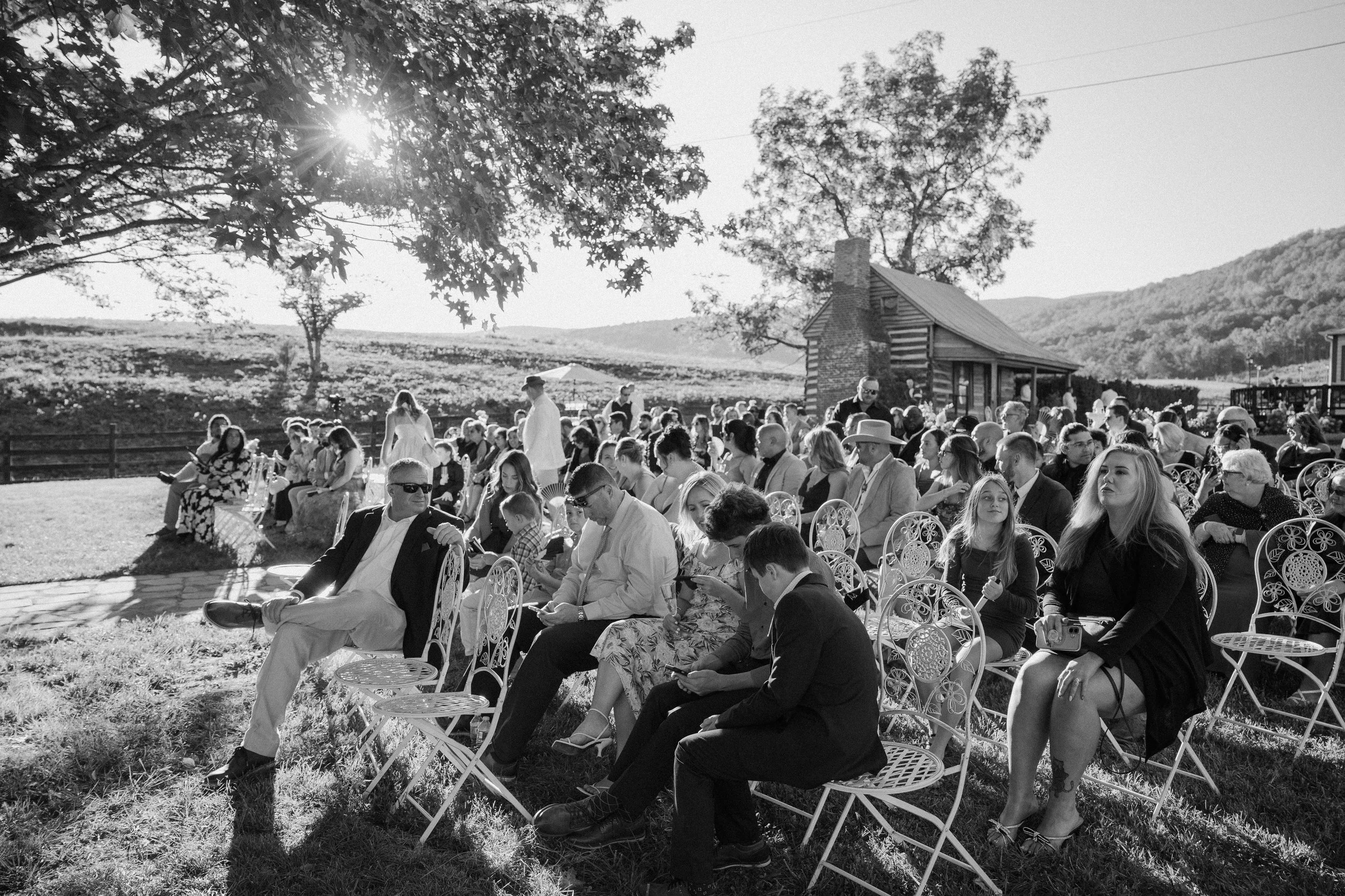 A large outdoor gathering of people seated on decorative chairs in a scenic rural setting with rolling hills, trees, and a rustic building, possibly a wedding or community event, during daylight.