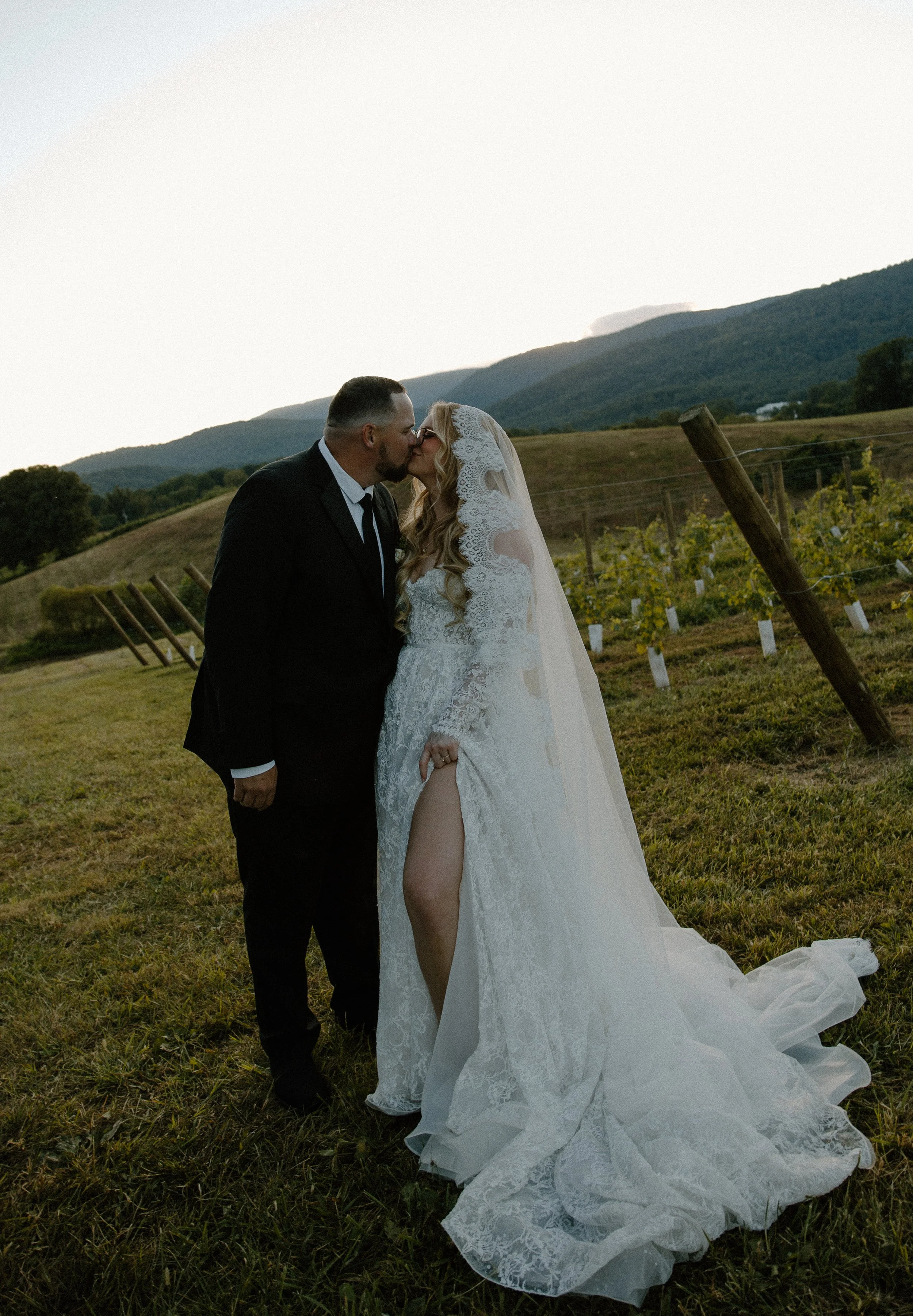 A newlywed couple kissing outdoors in a rural vineyard setting at sunset, with rolling hills in the background.