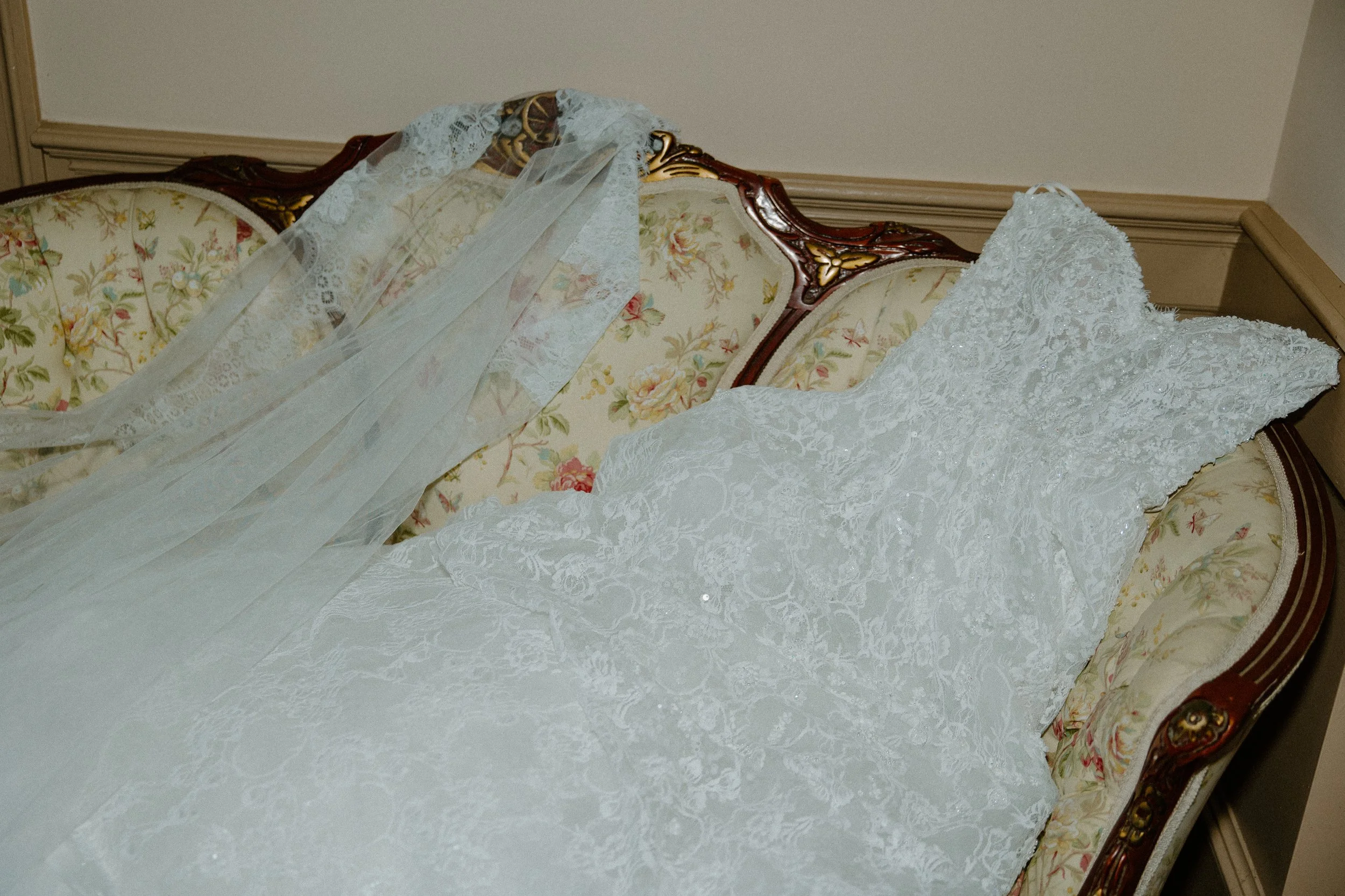 A floral upholstered vintage sofa with intricate wood carvings, lying on its side with a white lace wedding dress draped over its back and seat.