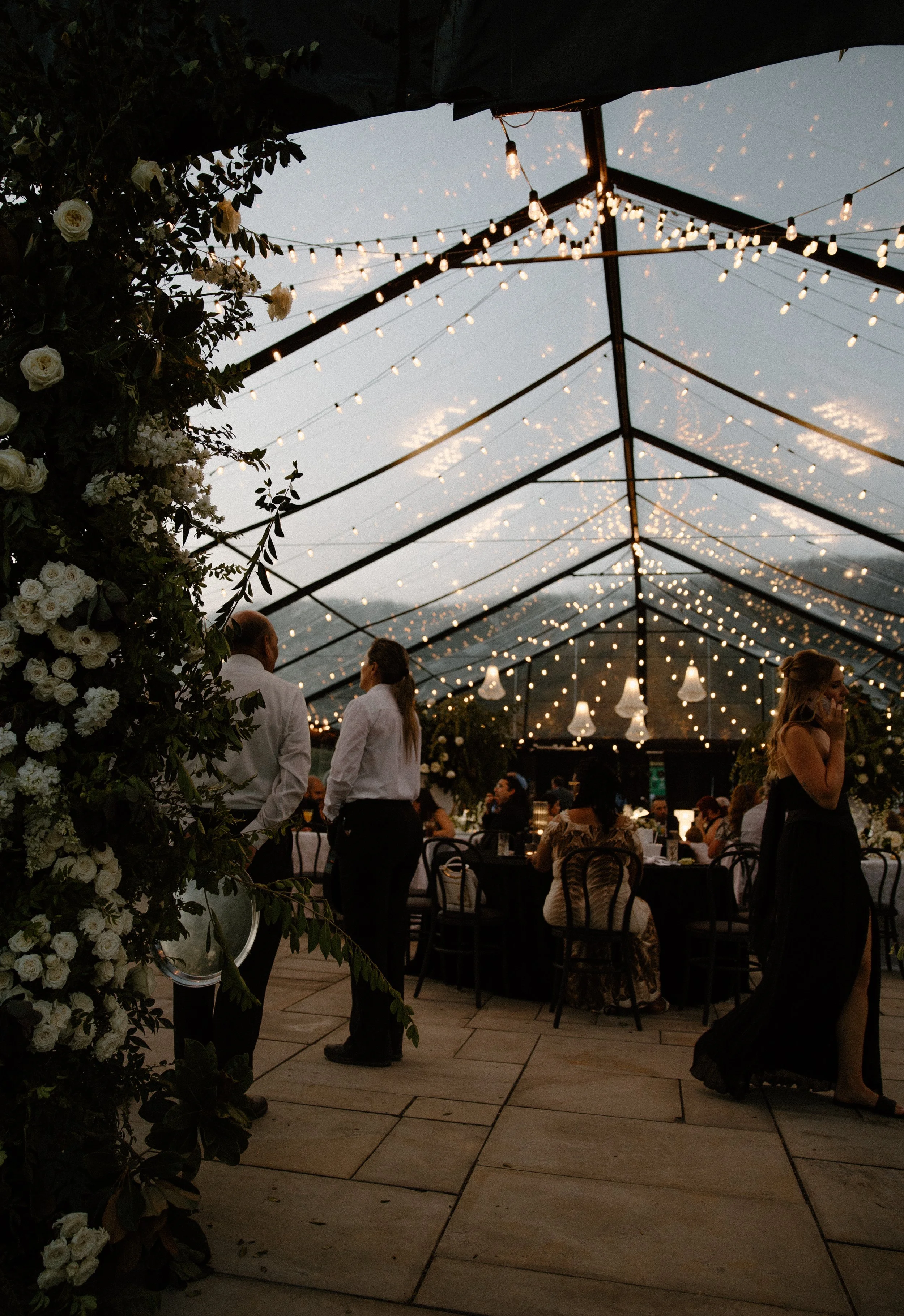 Outdoor event space at dusk with string lights, guests dressed semi-formally, and floral arrangements.