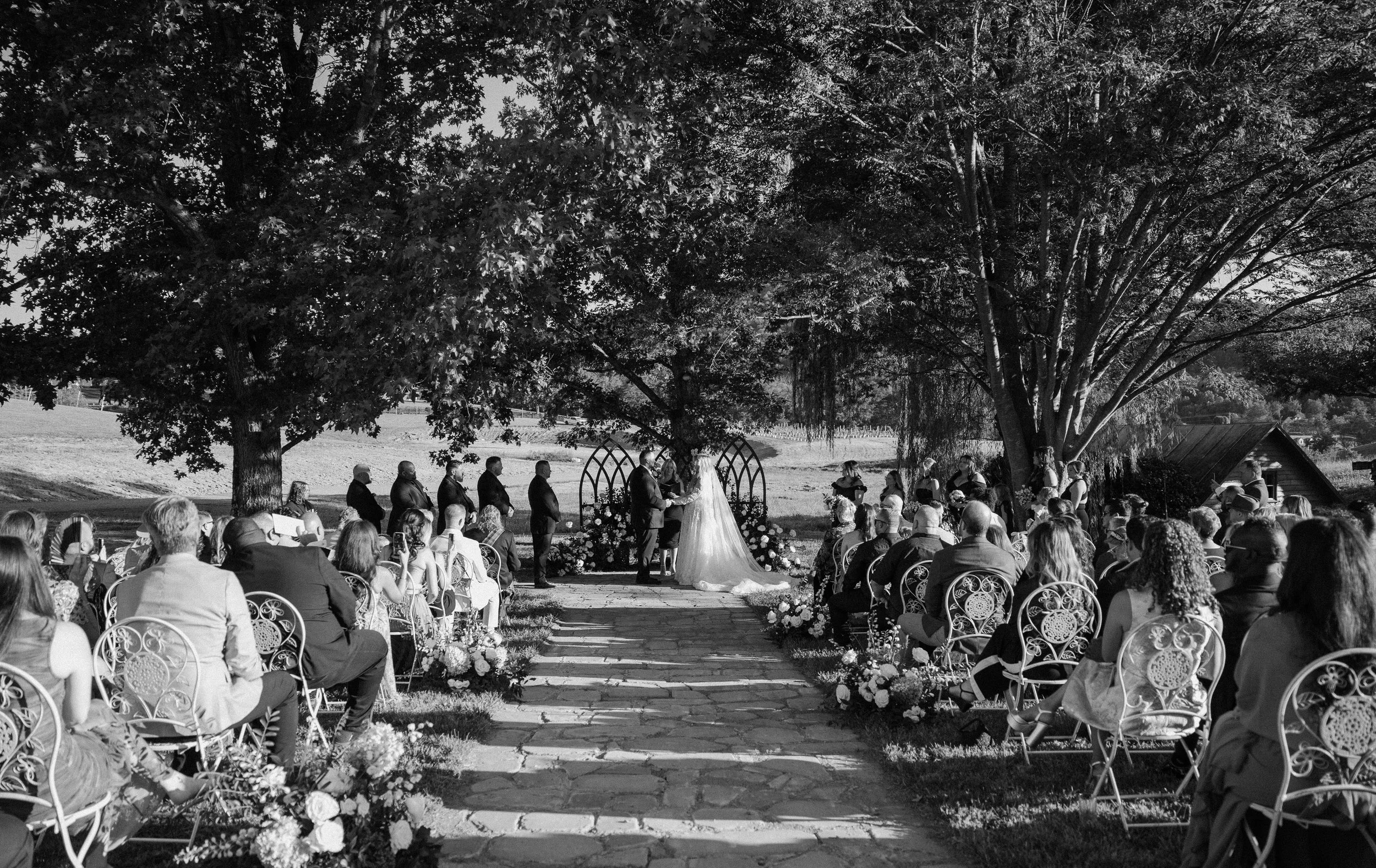 A black and white image of an outdoor wedding ceremony with guests seated on either side, facing a couple standing under a floral arch in front of a large tree. The bride is in a wedding gown with a veil, and the groom is in a suit. There is a scenic