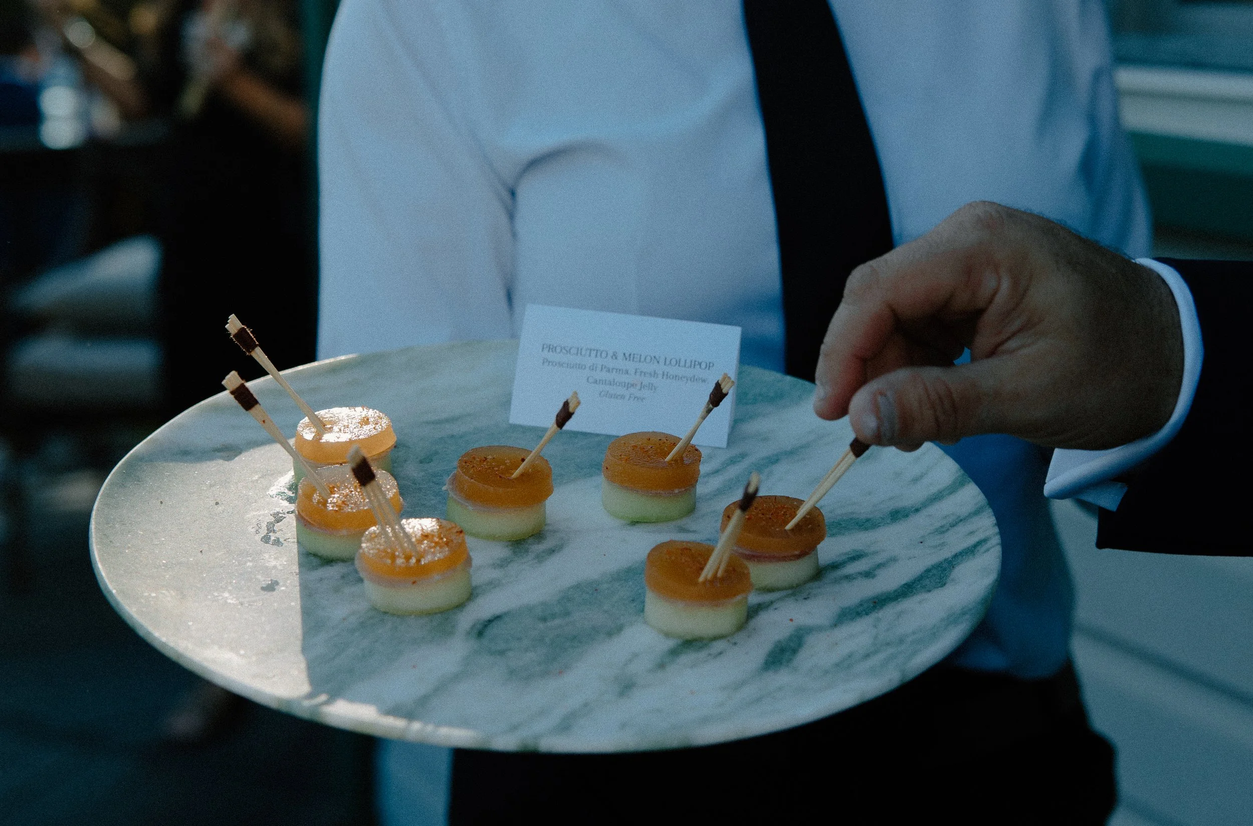A person in a formal suit is holding a large silver tray with small layered desserts topped with orange and white icing, garnished with small chocolate sticks.