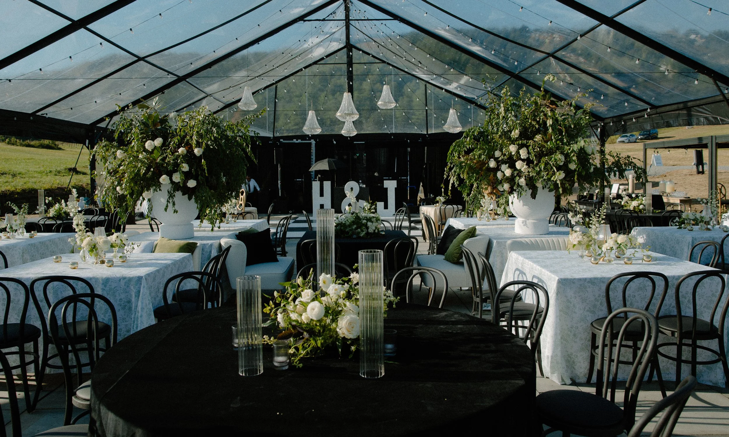 Outdoor wedding reception setup under a transparent tent with tables decorated with flowers and candles, hanging lights, and greenery surrounds.