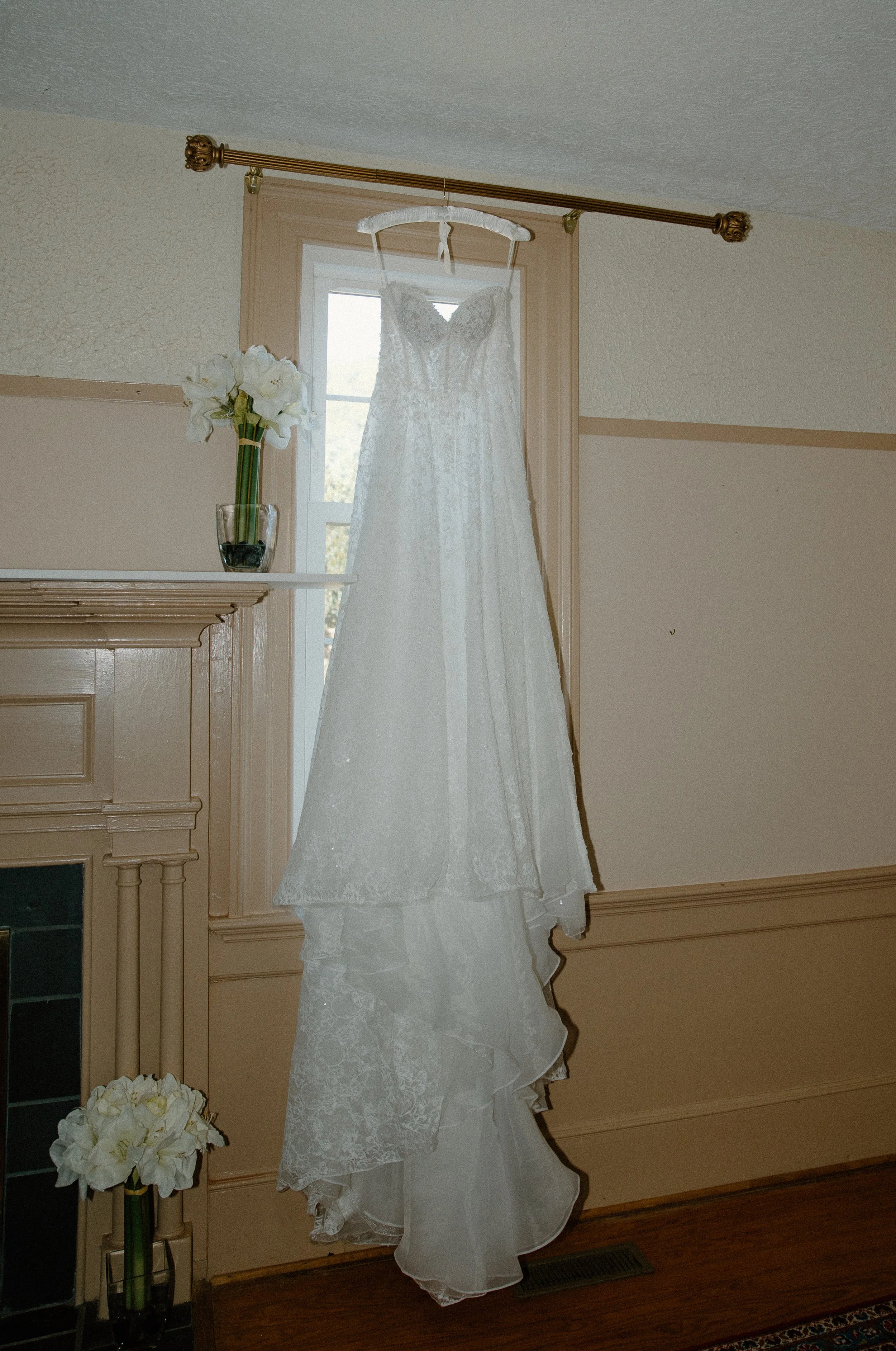 A white wedding dress hanging on a hook in front of a window, with a white flower arrangement on a table nearby, inside a room with beige walls and wooden trim.