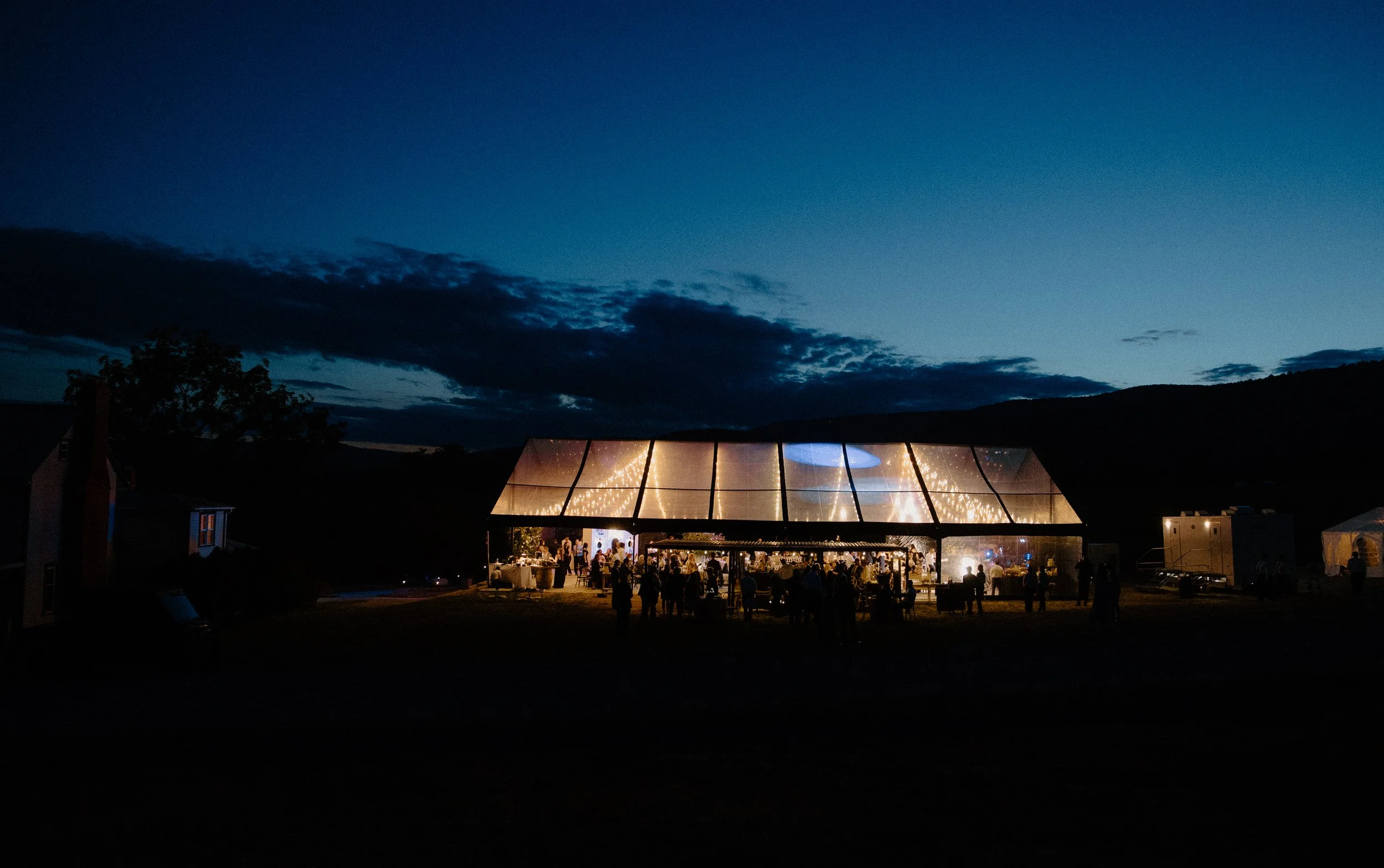 A nighttime outdoor event under a glass pavilion, with guests seated at tables and lights illuminating the structure, against a dark sky with clouds.