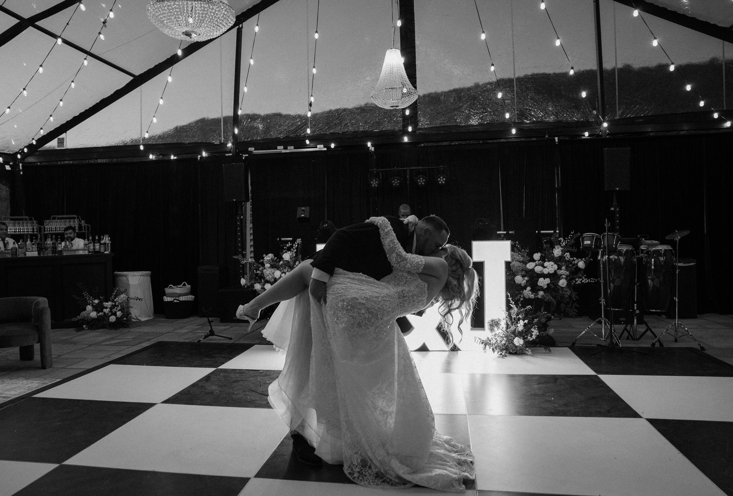 A bride and groom are dancing at their wedding reception inside a decorated venue with string lights and chandeliers. The bride is in a wedding gown, and the groom is in a suit. Their reflection is visible in the window behind them, and a band setup 