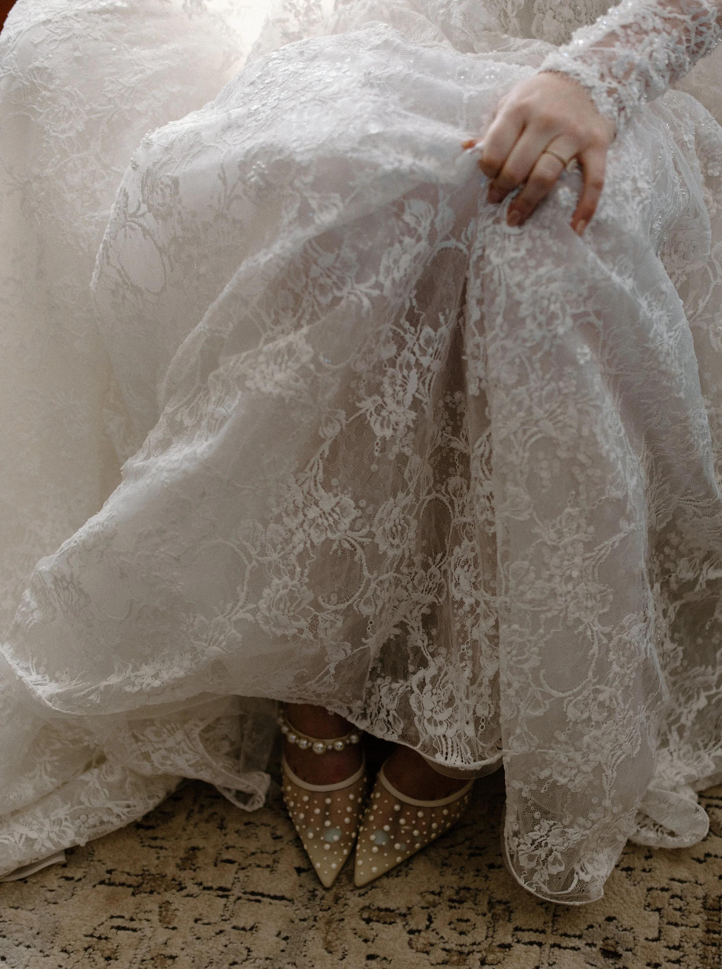 The image shows a person dressed in a white lace dress, possibly a wedding gown, with a focus on their feet. They are wearing beige shoes with white polka dots and a pearl ankle bracelet. The person is holding up the skirt of the dress, revealing the shoes and part of their leg.