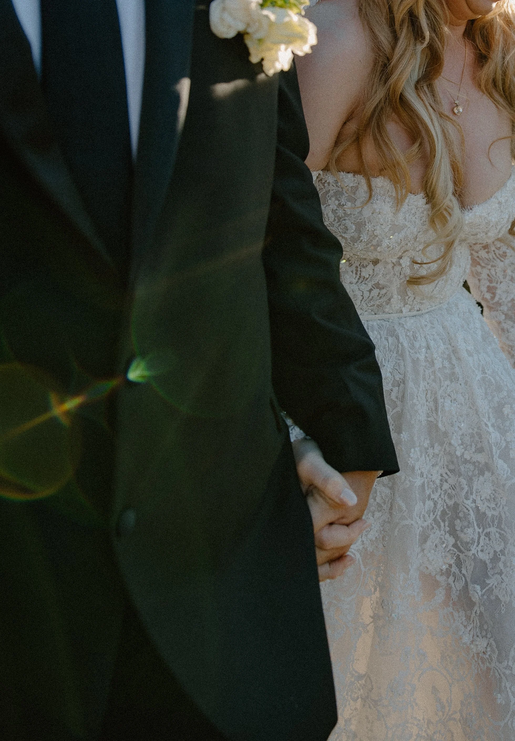 Close-up of a couple holding hands, the man in a dark suit and the woman in a lace wedding dress with a pearl necklace, outdoors during the daytime.
