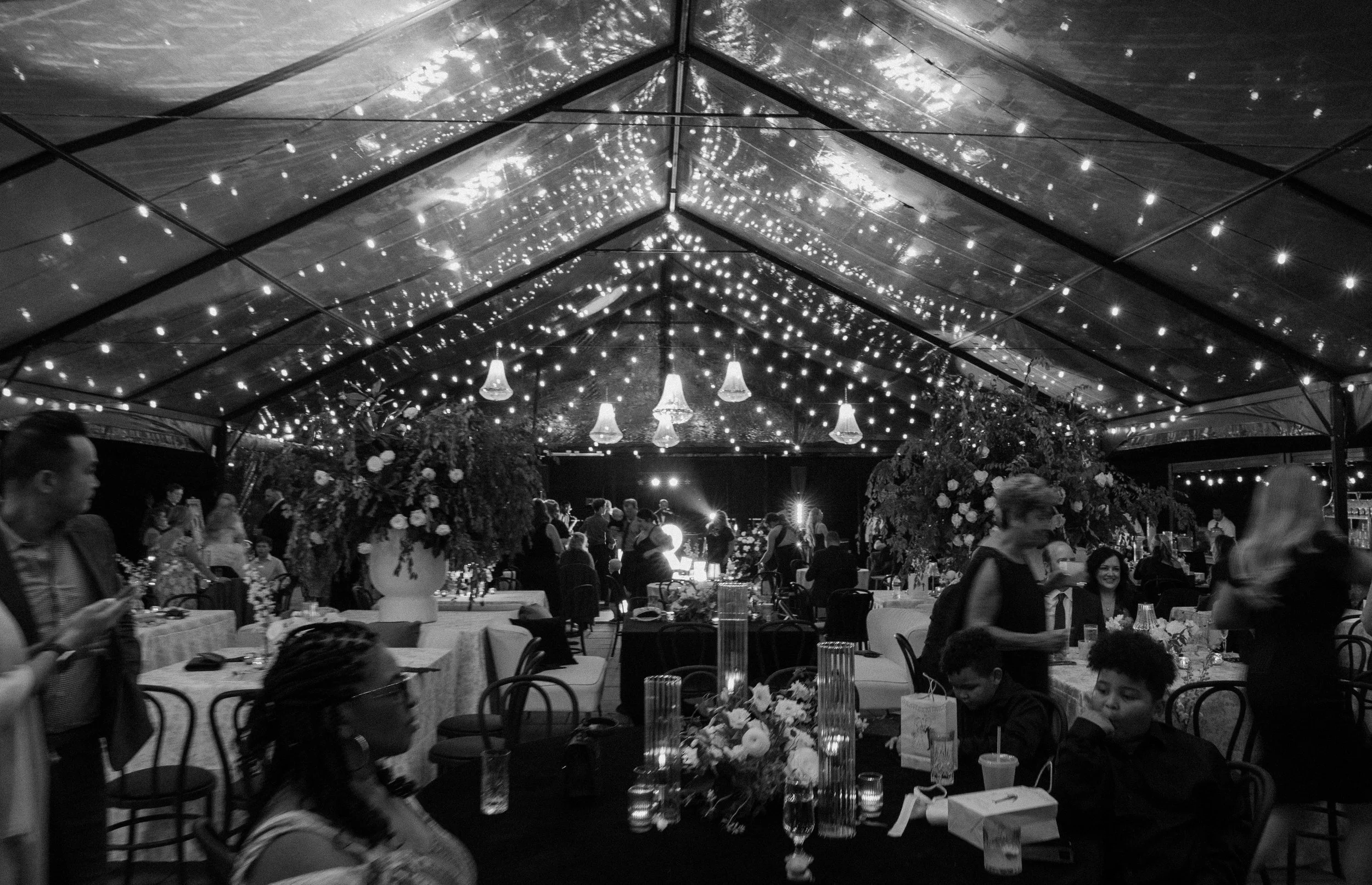 A black and white photo of a decorated event venue with string lights on the ceiling, tables set with flowers and candles, and guests seated and walking around.