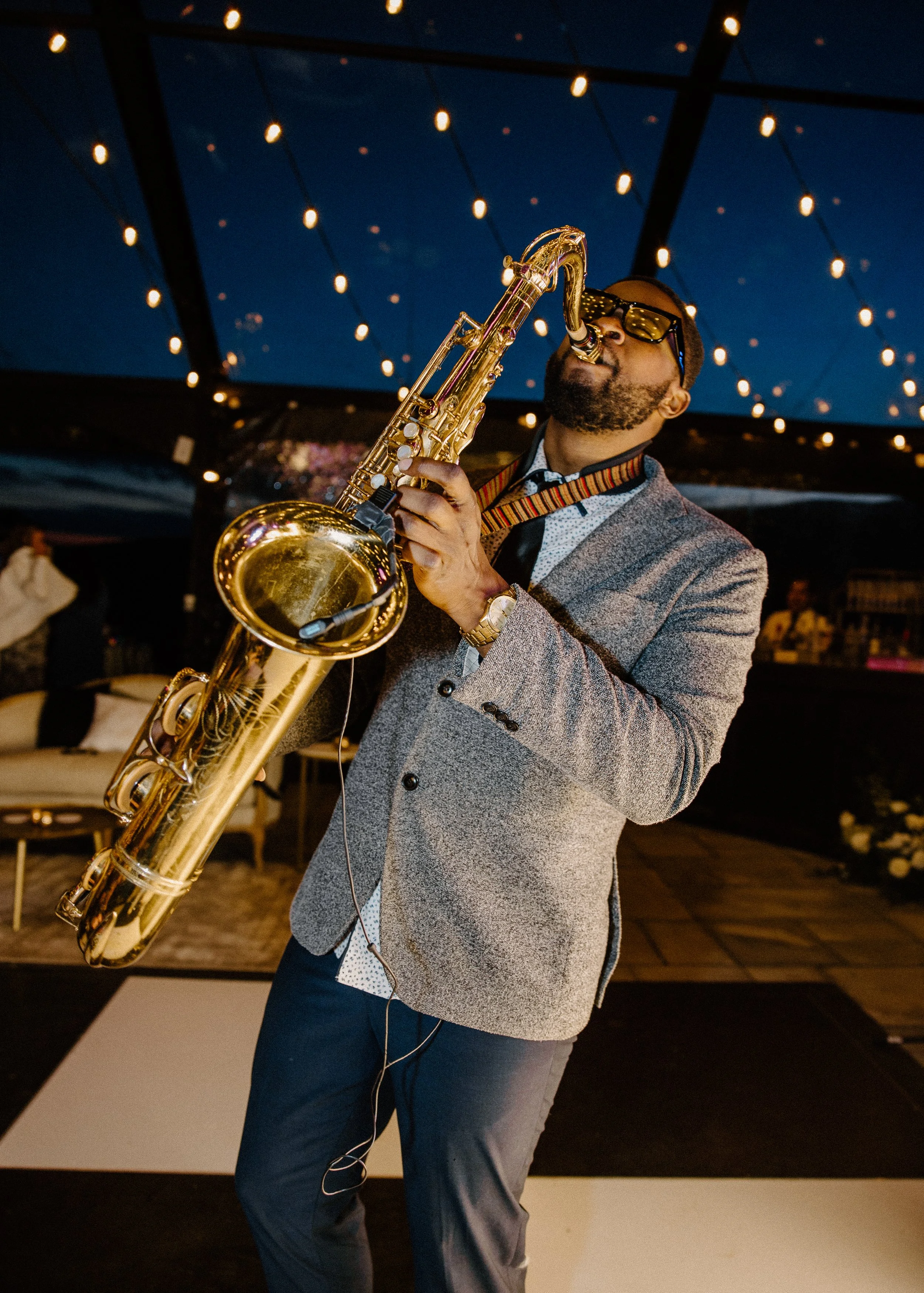 A man in a gray blazer, white shirt, and dark pants playing a gold saxophone at an outdoor event during the evening, with string lights overhead.