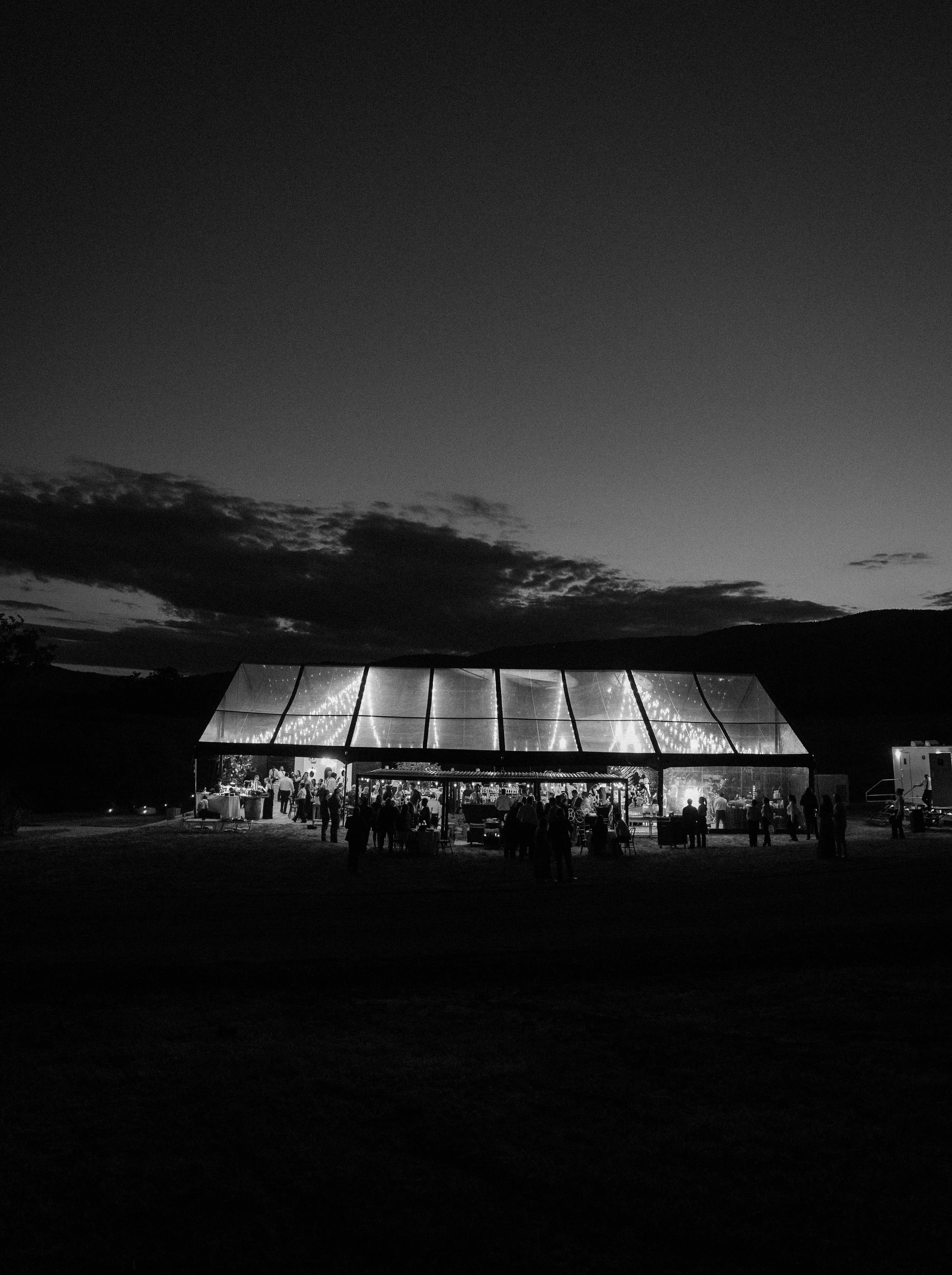 Black and white photo of an outdoor gathering at night in front of a modern glass building with illuminated interior, surrounded by a natural landscape with clouds and hills.
