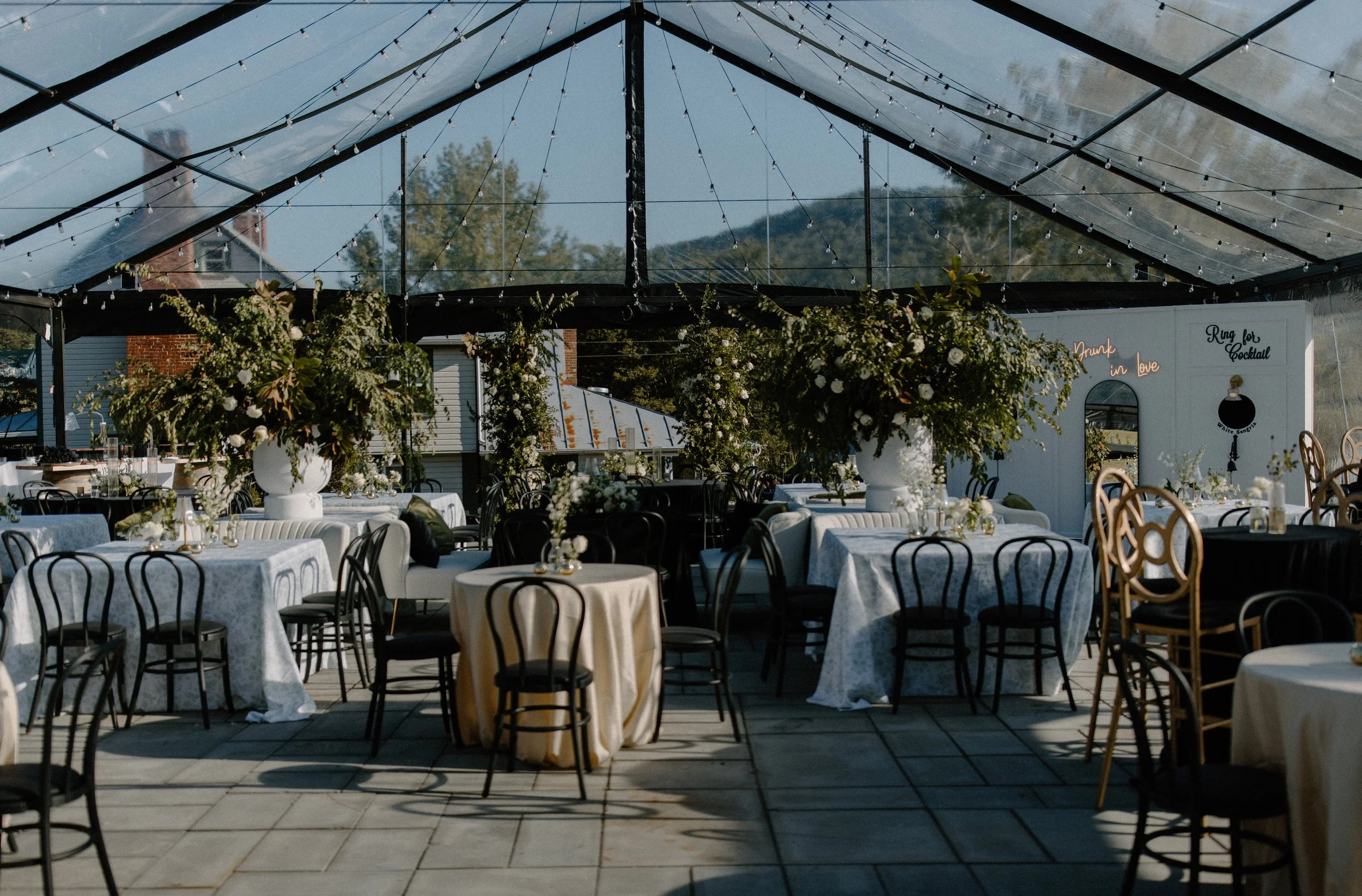 Outdoor wedding reception setup with round tables covered in white tablecloths and black chairs, floral centerpieces, string lights, and a white backdrop with a sign reading 'Drink in Love' and 'Run for Cocktail' in neon lights.