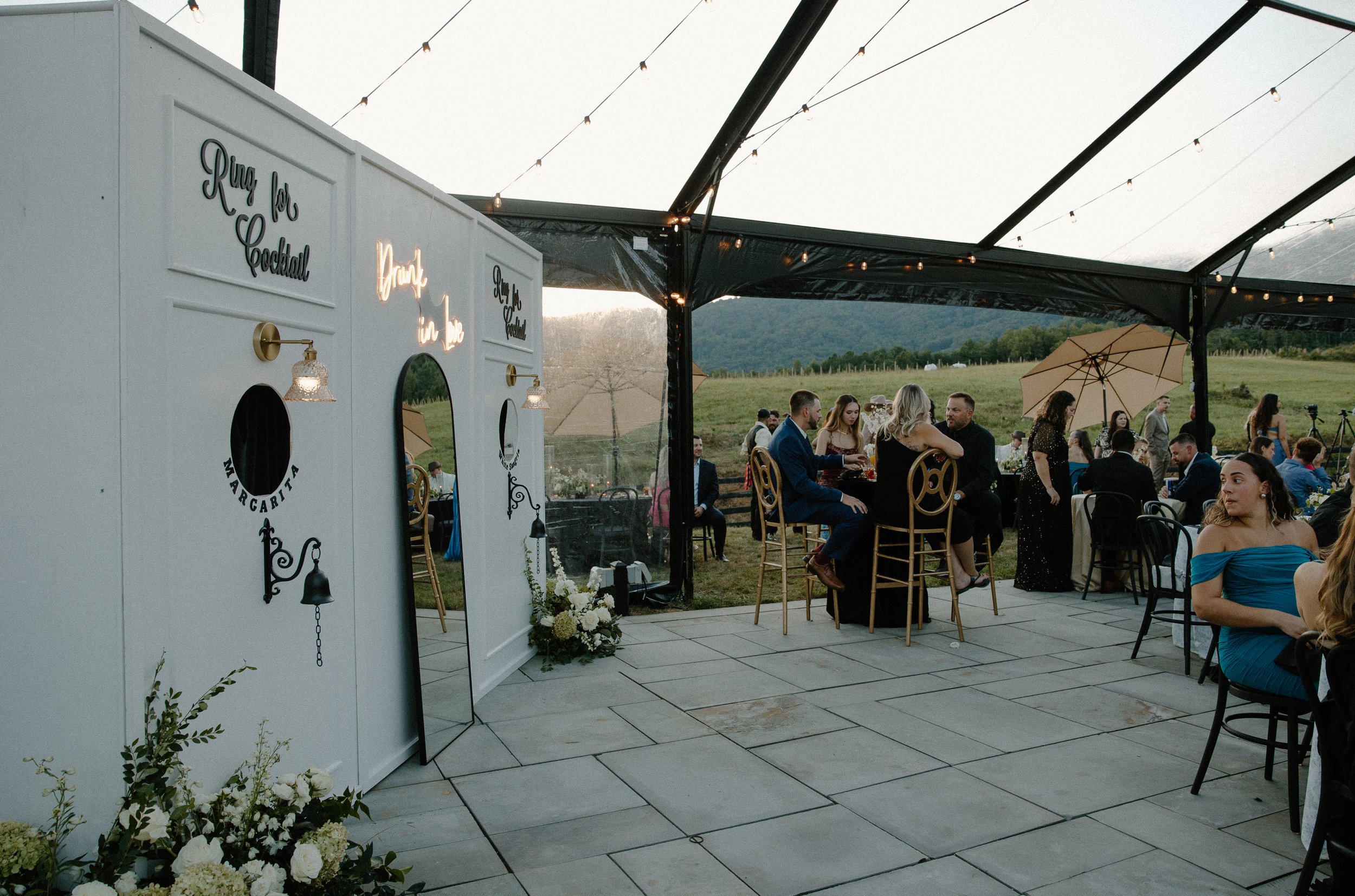 Guests seated at tables during an outdoor wedding reception under a black canopy with string lights. A white photo booth with hanging signs and floral decorations is visible on the left. The background features green hills and a partly cloudy sky.